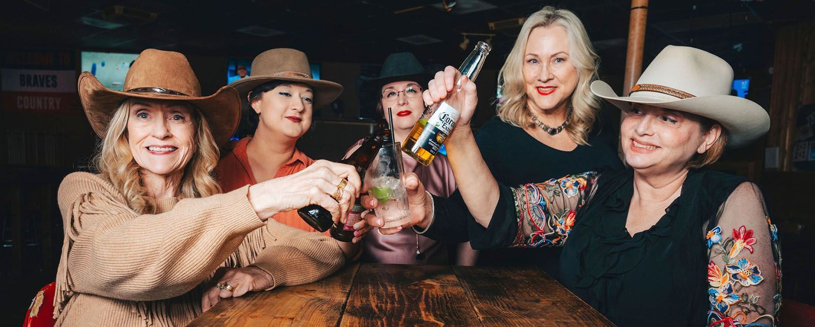 Five women in cowboy hats clink bottles at a bar