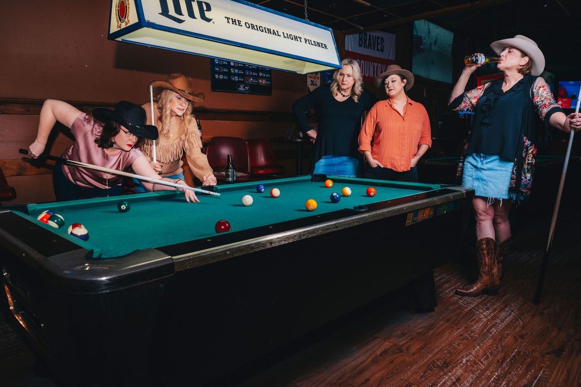 People playing pool in a dim bar, with a billiards table and hanging sign overhead.