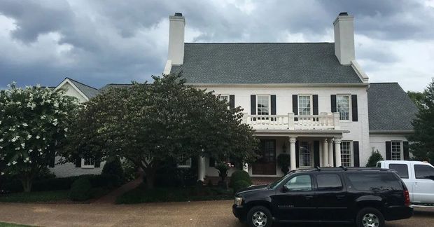 White two-story house with porch, trees, and parked vehicles in front under a cloudy sky