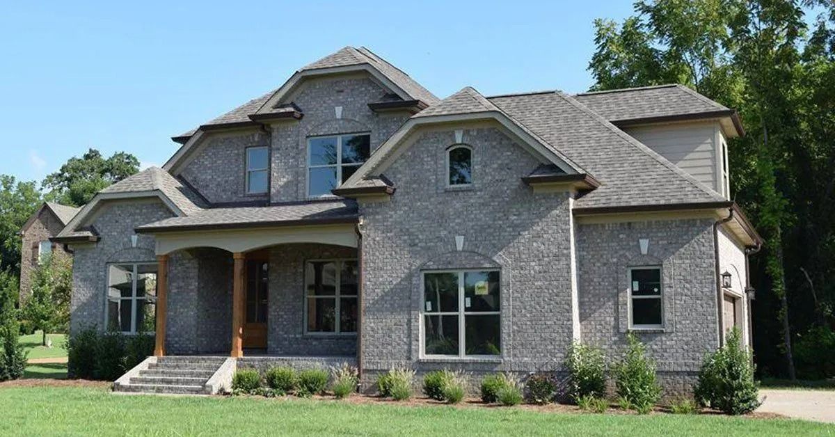 Large gray stone suburban house with a front porch and gabled roof, surrounded by green lawn and trees
