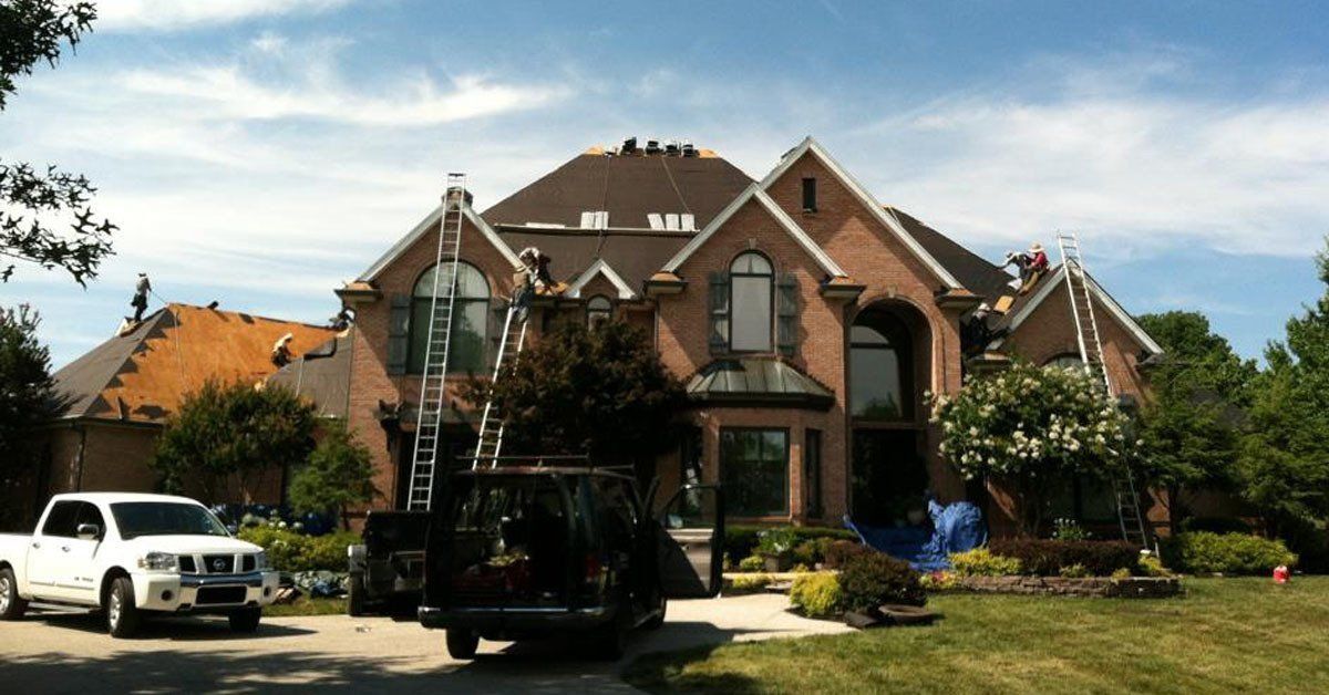 Large brick house with a steep roof, front porch, and parked vehicles in a landscaped yard.