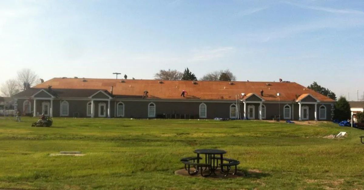 Long brick building with a red roof behind a grassy field and picnic table under a clear sky
