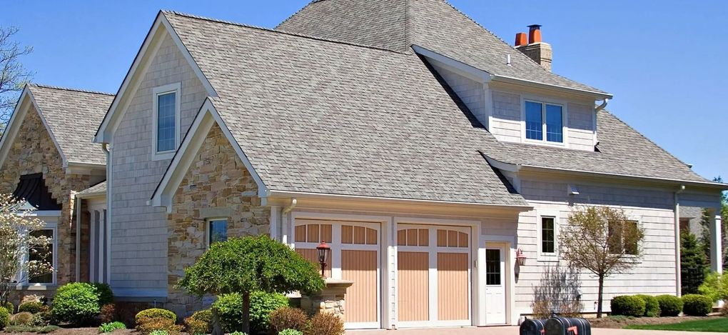 Two-story stone house with a gray shingled roof, white trim, and a front porch surrounded by landscaping