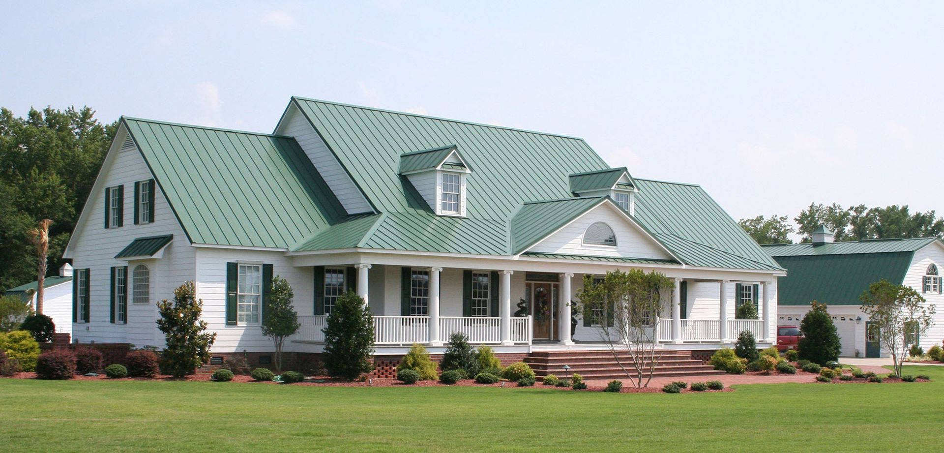 White farmhouse with green roofs and a wraparound porch on a landscaped lawn