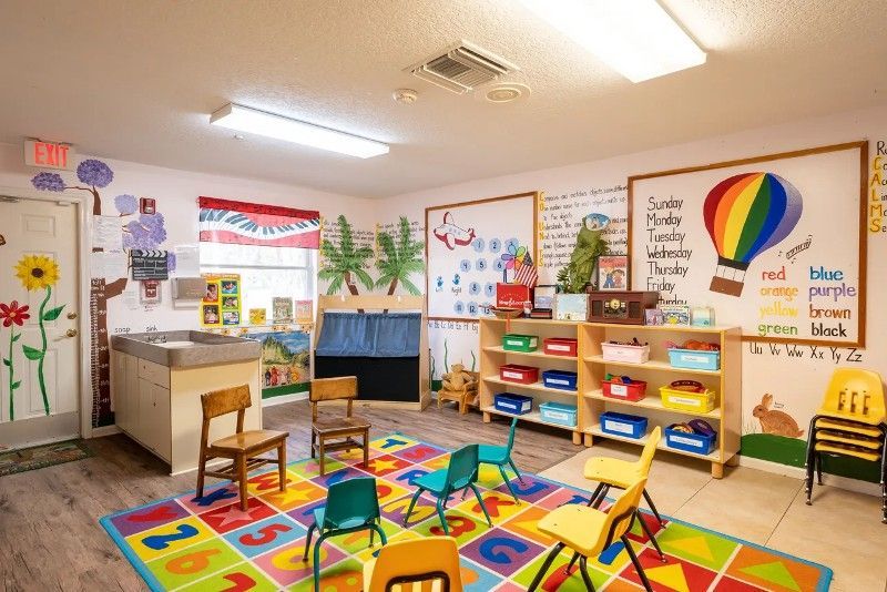 A classroom with a colorful rug , chairs , shelves and a changing table.