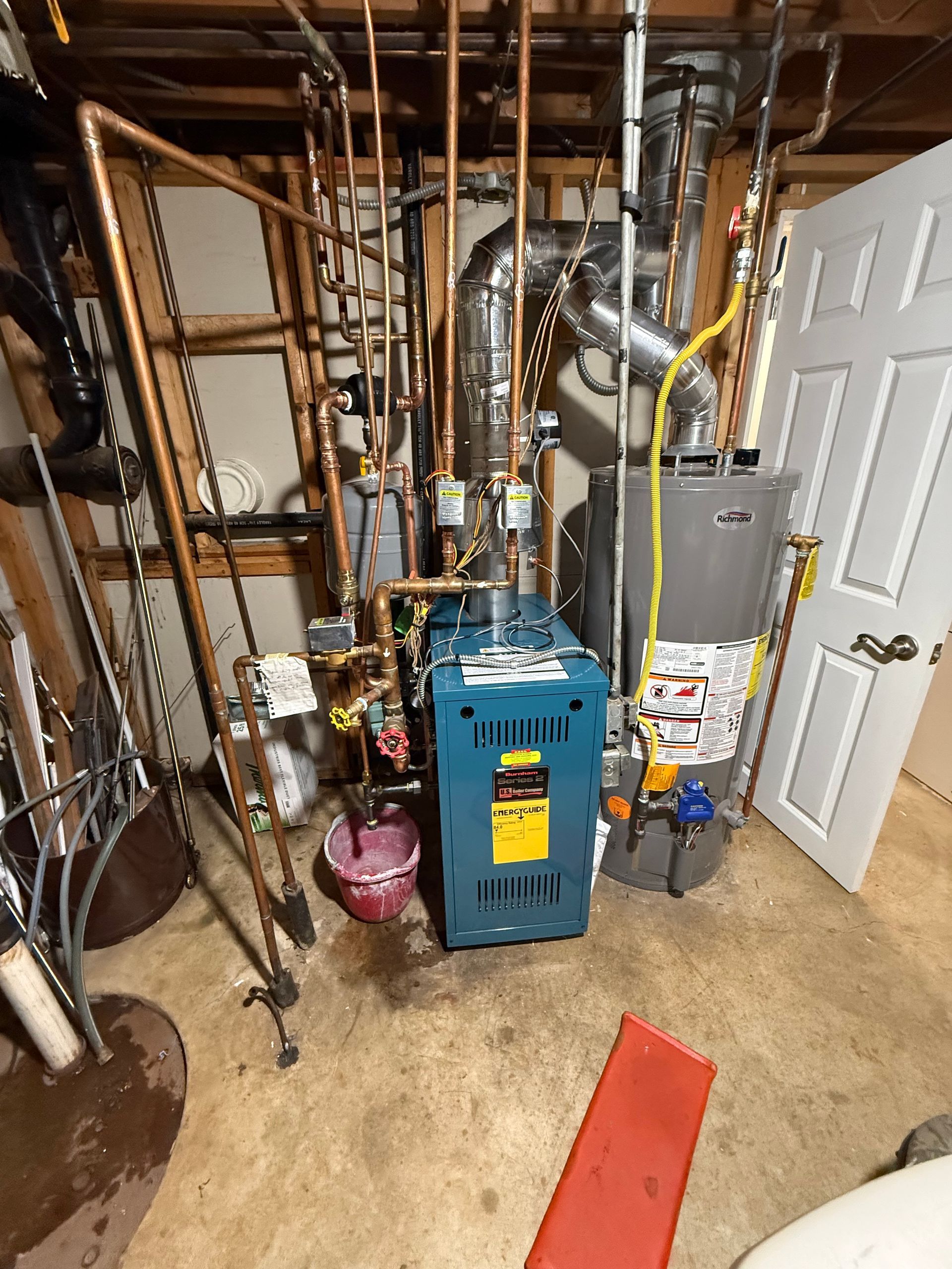 A utility room containing a blue boiler, a gray water heater, copper pipes, and a red bucket on a concrete floor.