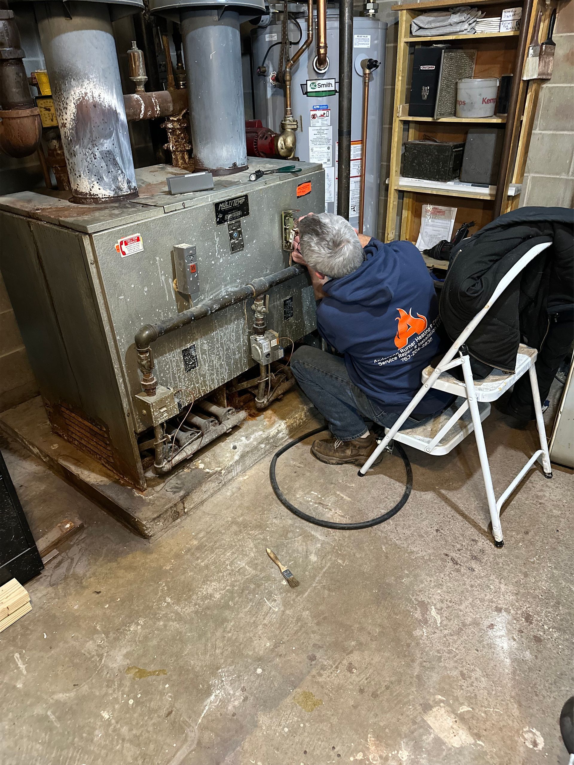 A technician wearing a navy hoodie kneels on a white step stool, working on a large, metal industrial boiler.