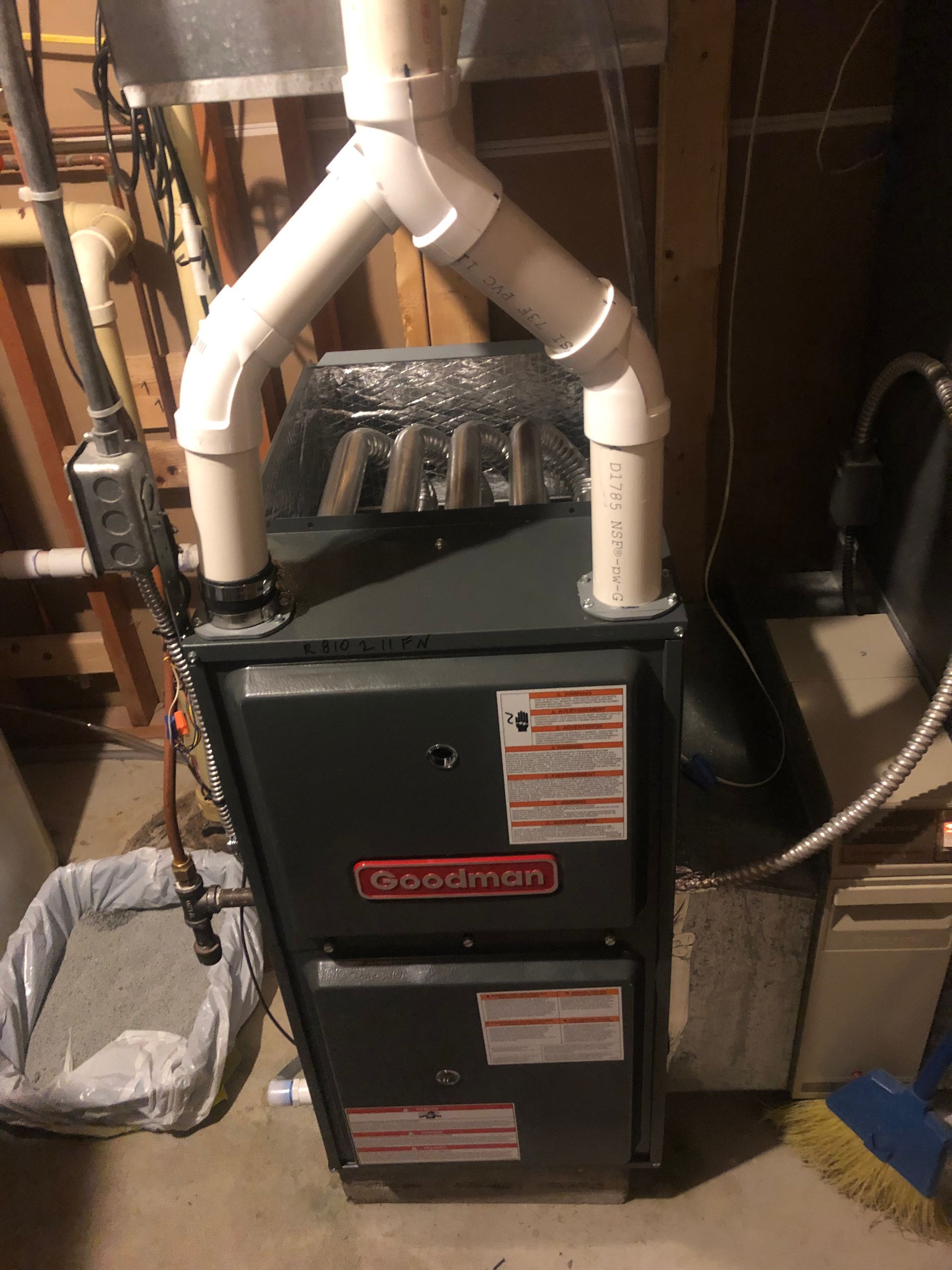 A grey Goodman furnace in a basement with white PVC exhaust piping connected to the top.