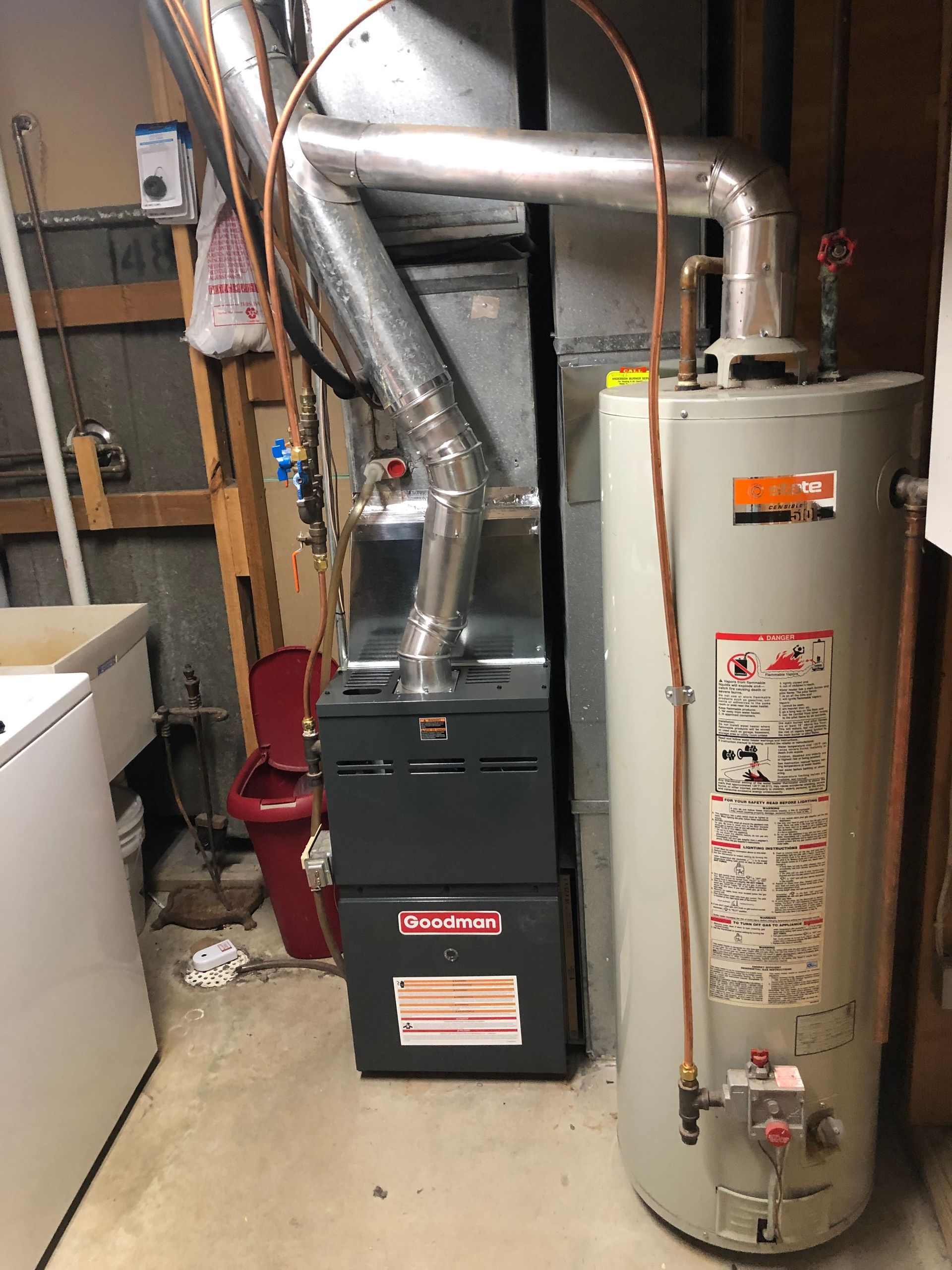 A grey water heater and a dark furnace stand side-by-side in a basement, connected by metal venting pipes.
