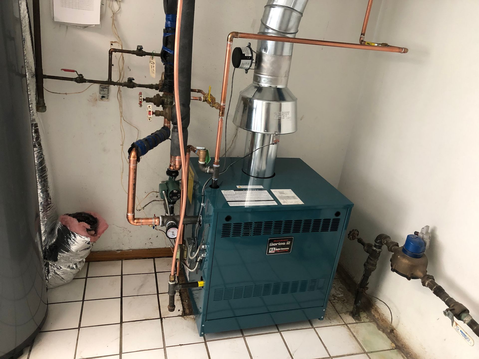 A teal residential boiler in a tiled room, connected to copper piping and a silver metal exhaust flue.