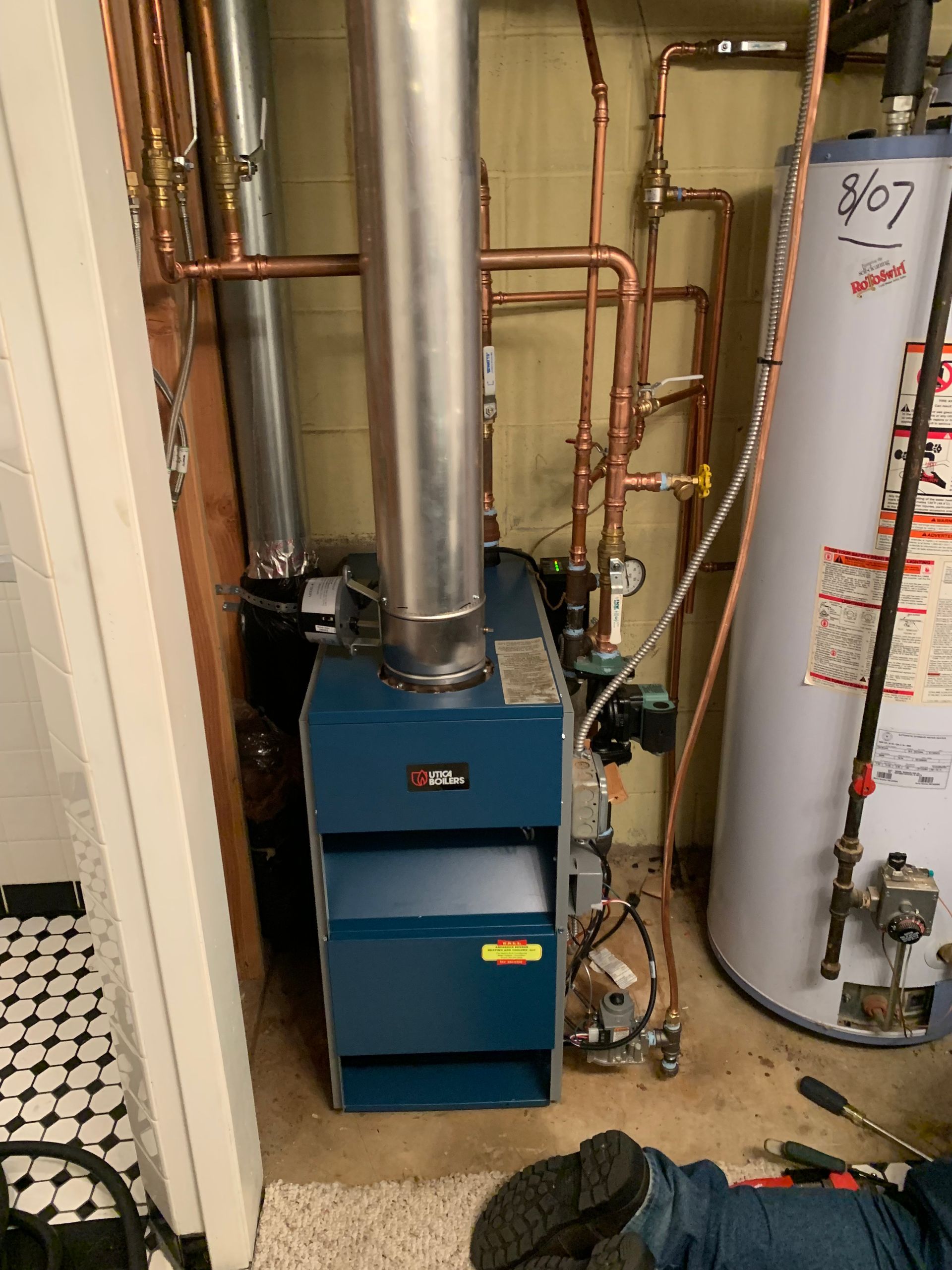A blue gas boiler for a heating system stands in a utility closet next to a water heater with copper piping above.