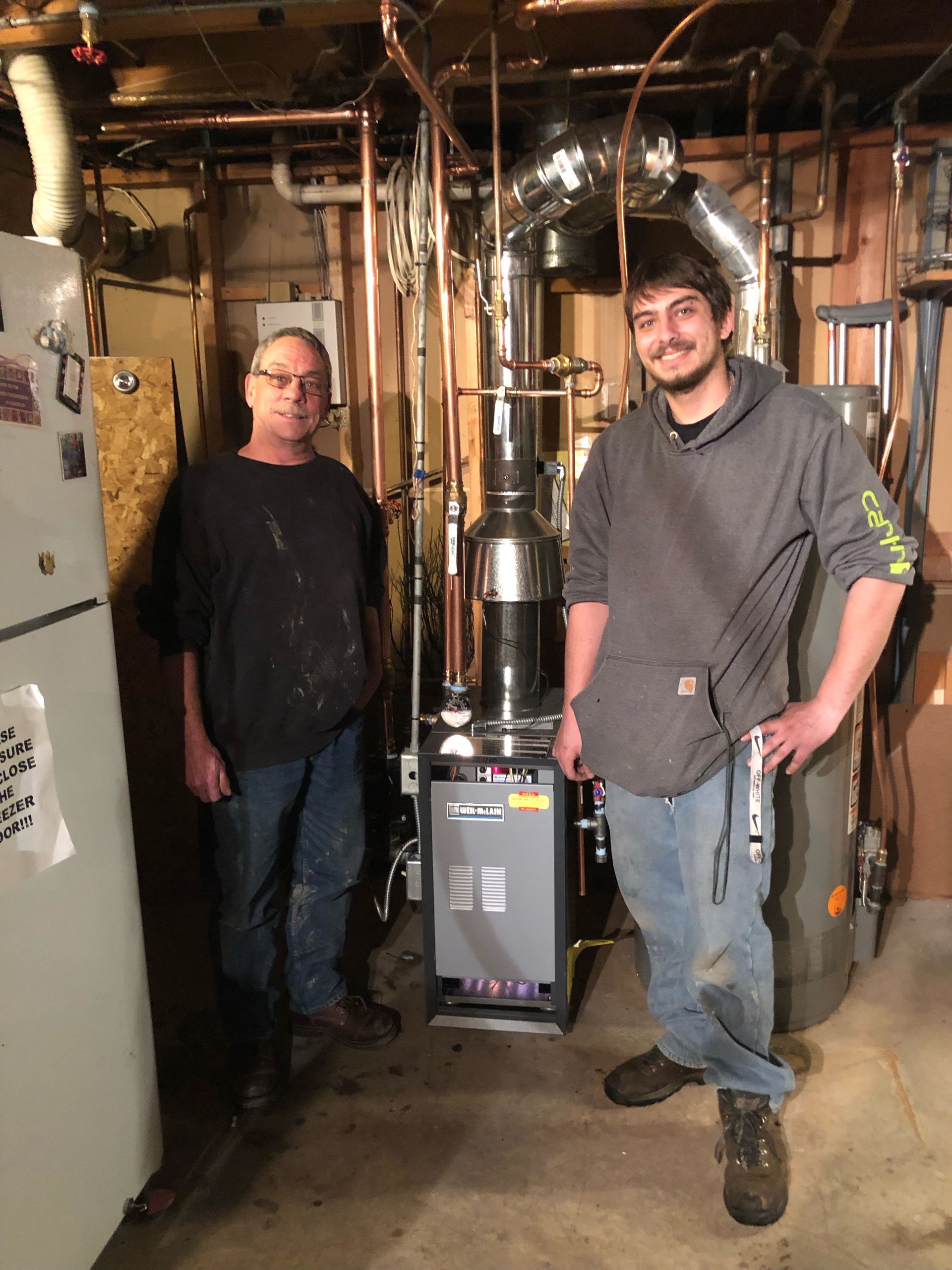 Two people stand in a basement beside a newly installed HVAC boiler unit surrounded by copper plumbing.