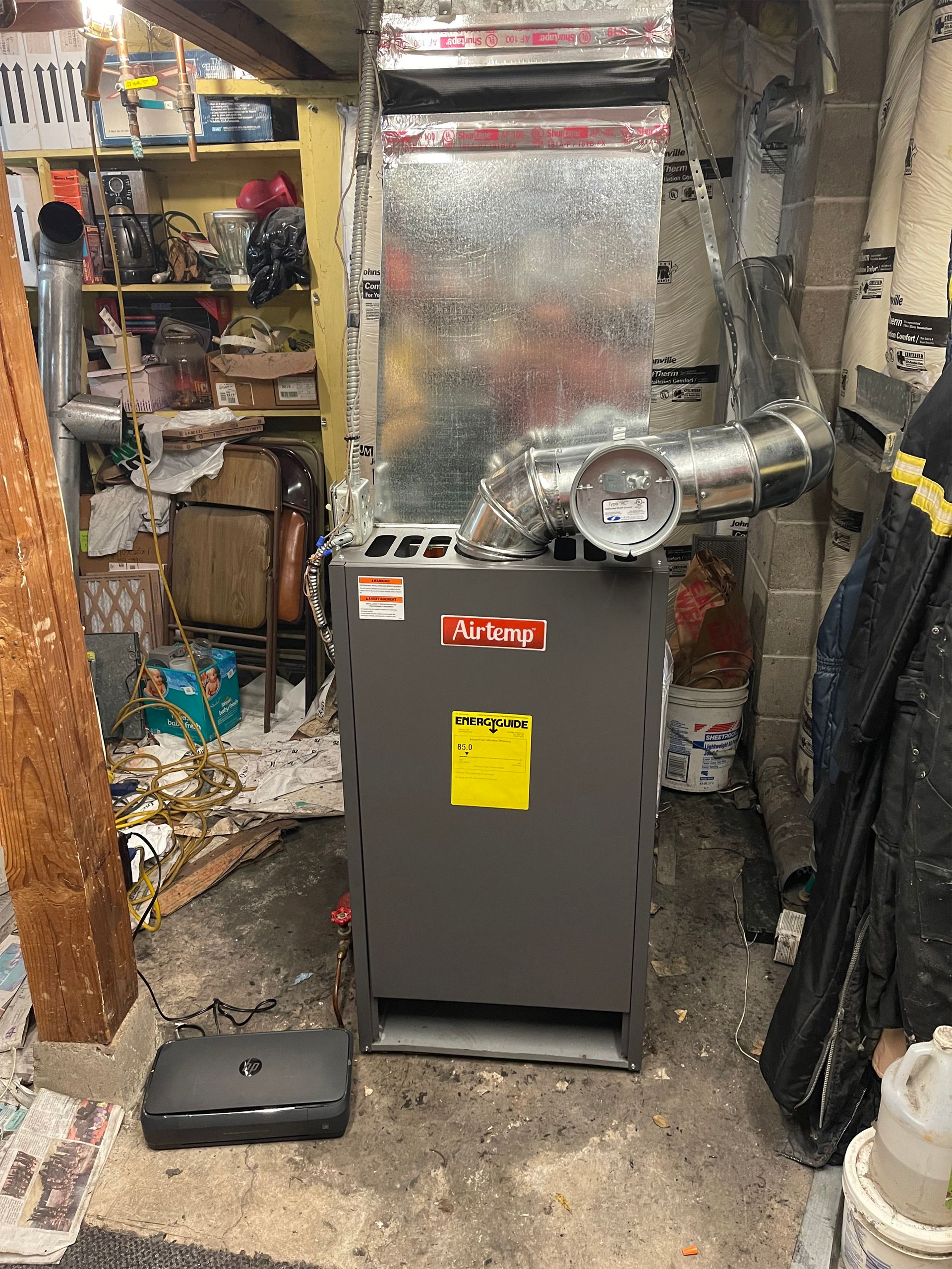 A grey Armstrong Air furnace stands in a cluttered basement with metal ductwork attached to the top.