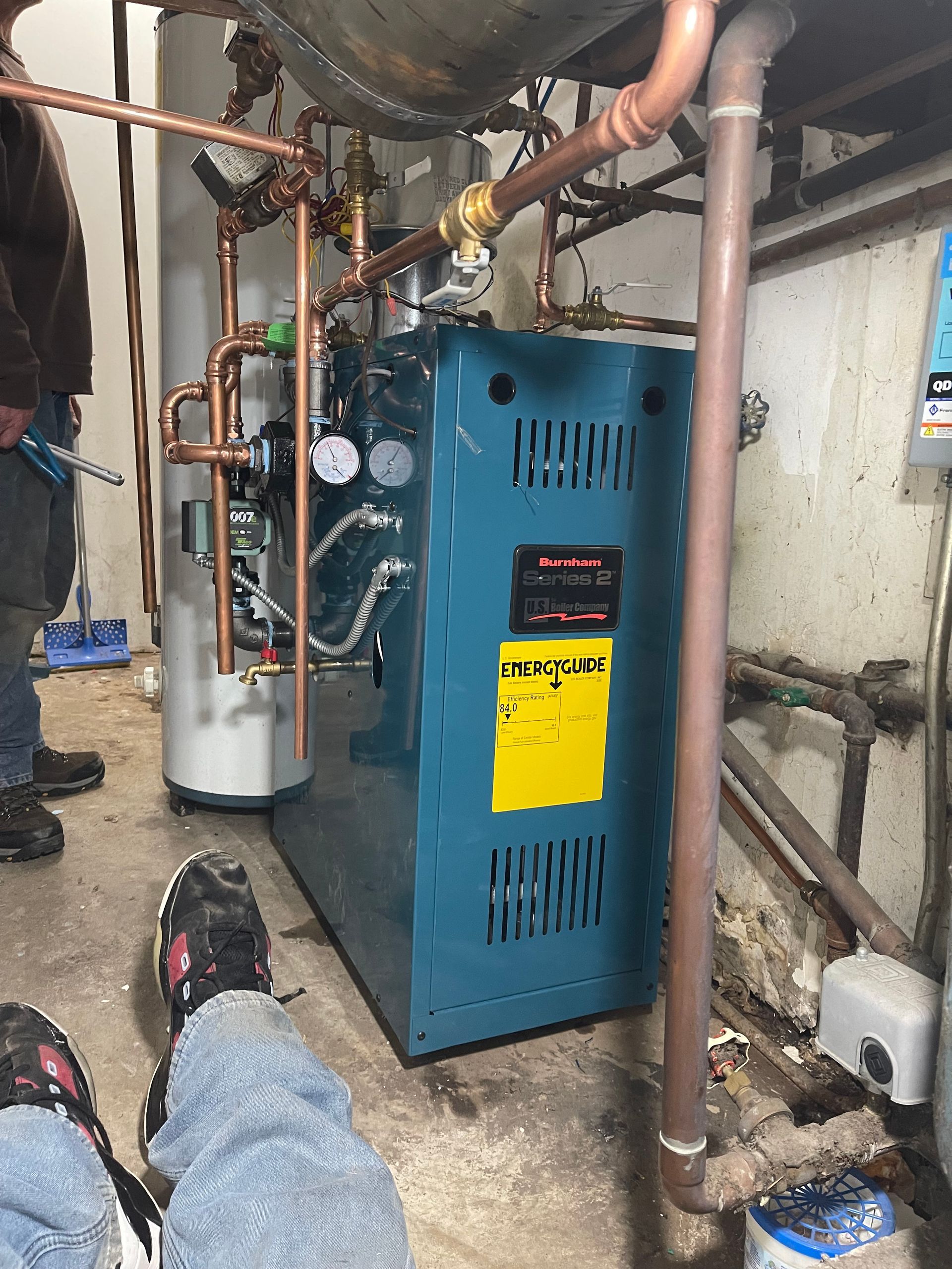A technician working on a blue boiler in a basement with various copper pipes and equipment visible.
