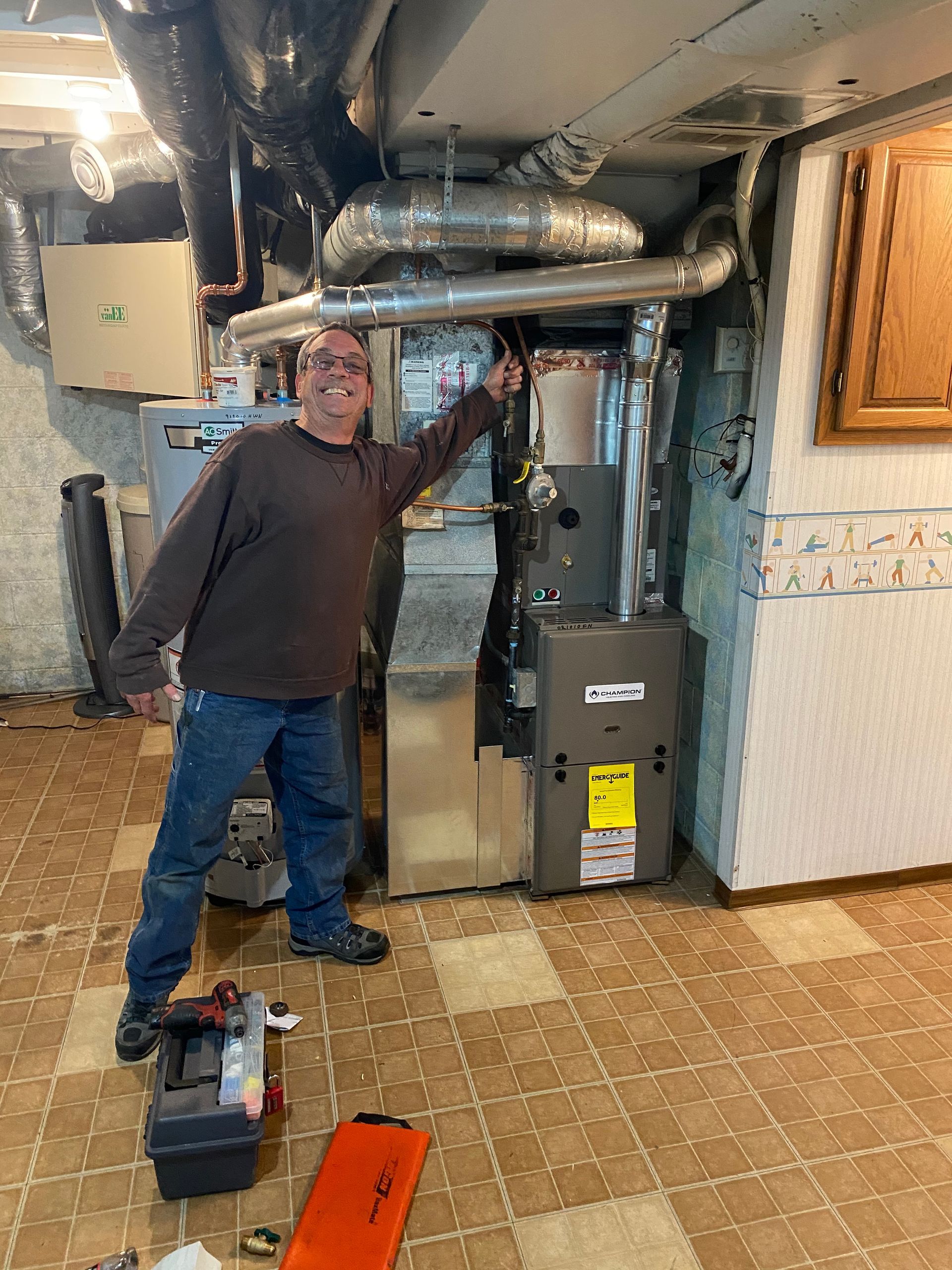 A smiling person stands in a basement next to a newly installed grey furnace, pointing toward the ceiling ductwork.