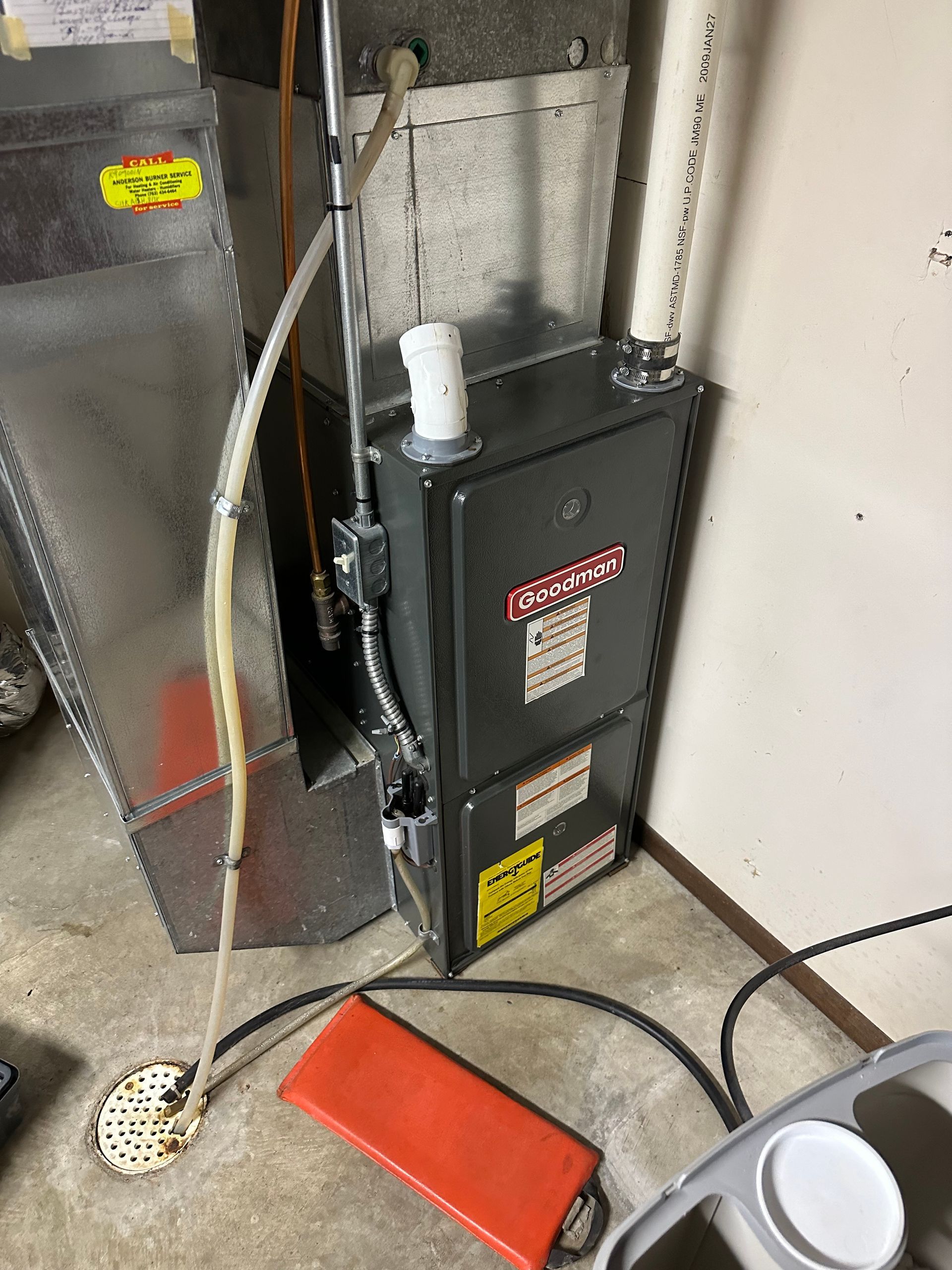 A grey HVAC furnace unit with white PVC piping, standing next to a metal duct in a basement setting.