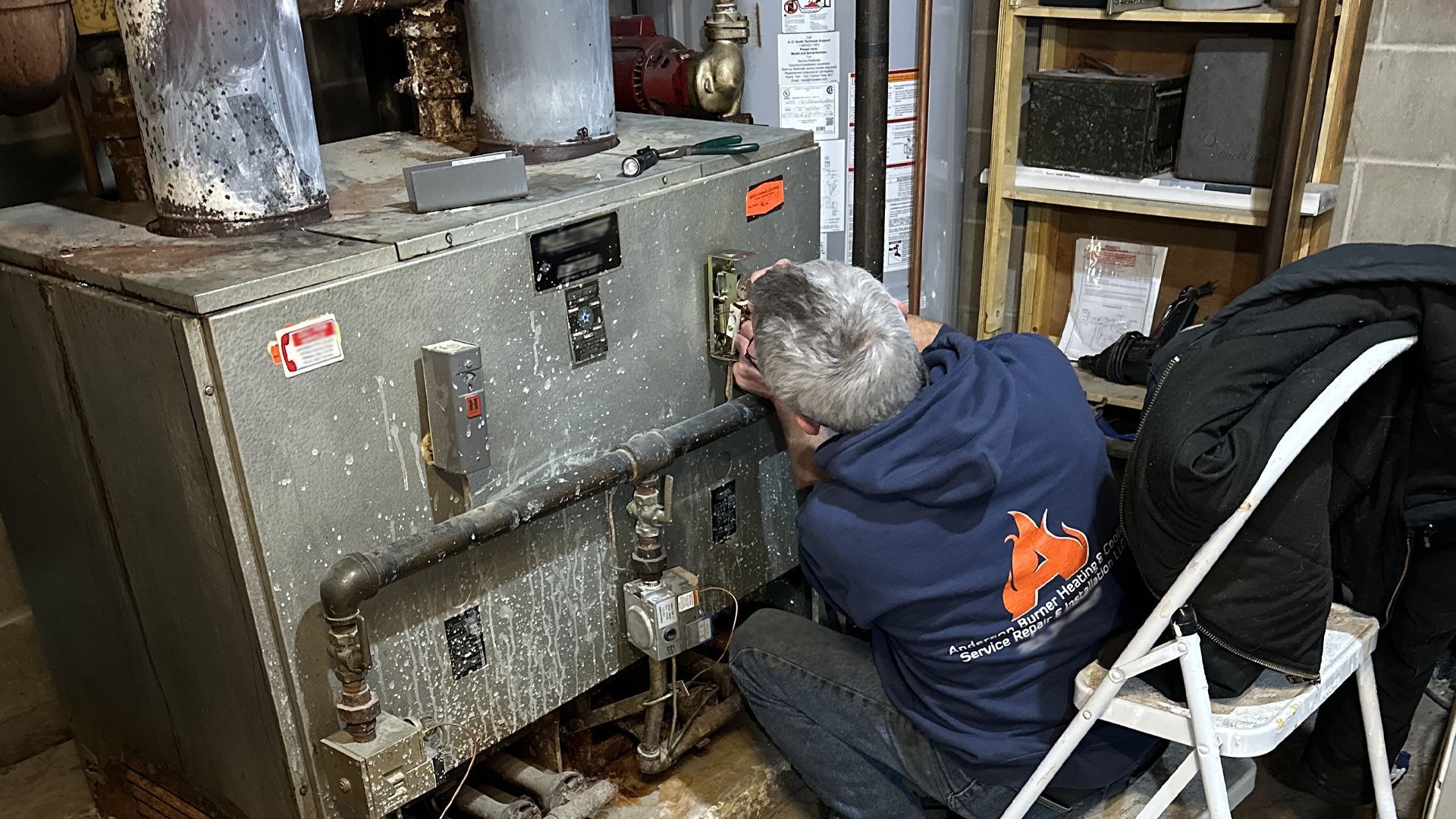 A technician wearing a navy blue sweatshirt services an industrial boiler in a utility room.