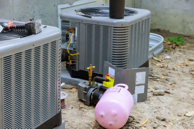 Two HVAC units sitting outdoors with a vacuum pump and a pink refrigerant cylinder connected to one for maintenance.