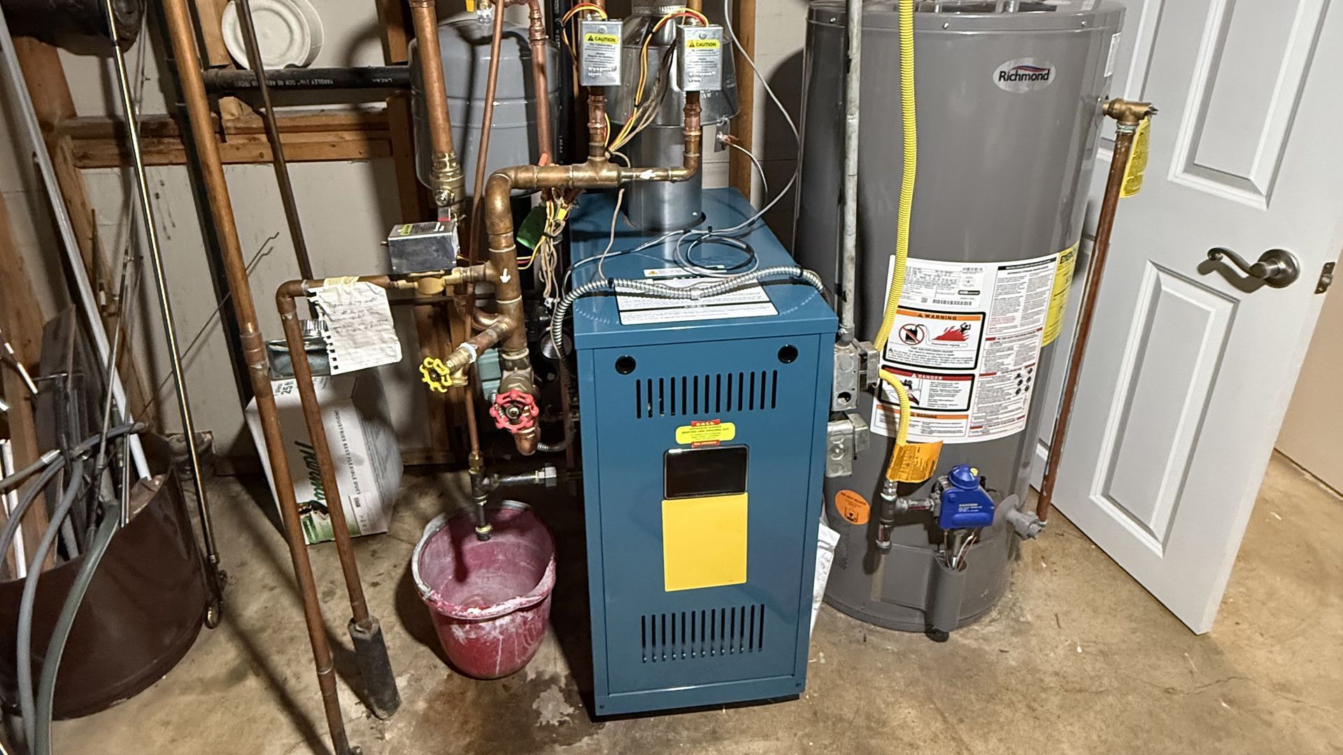A residential boiler and water heater in a basement, with a red bucket catching water dripping from copper pipes.