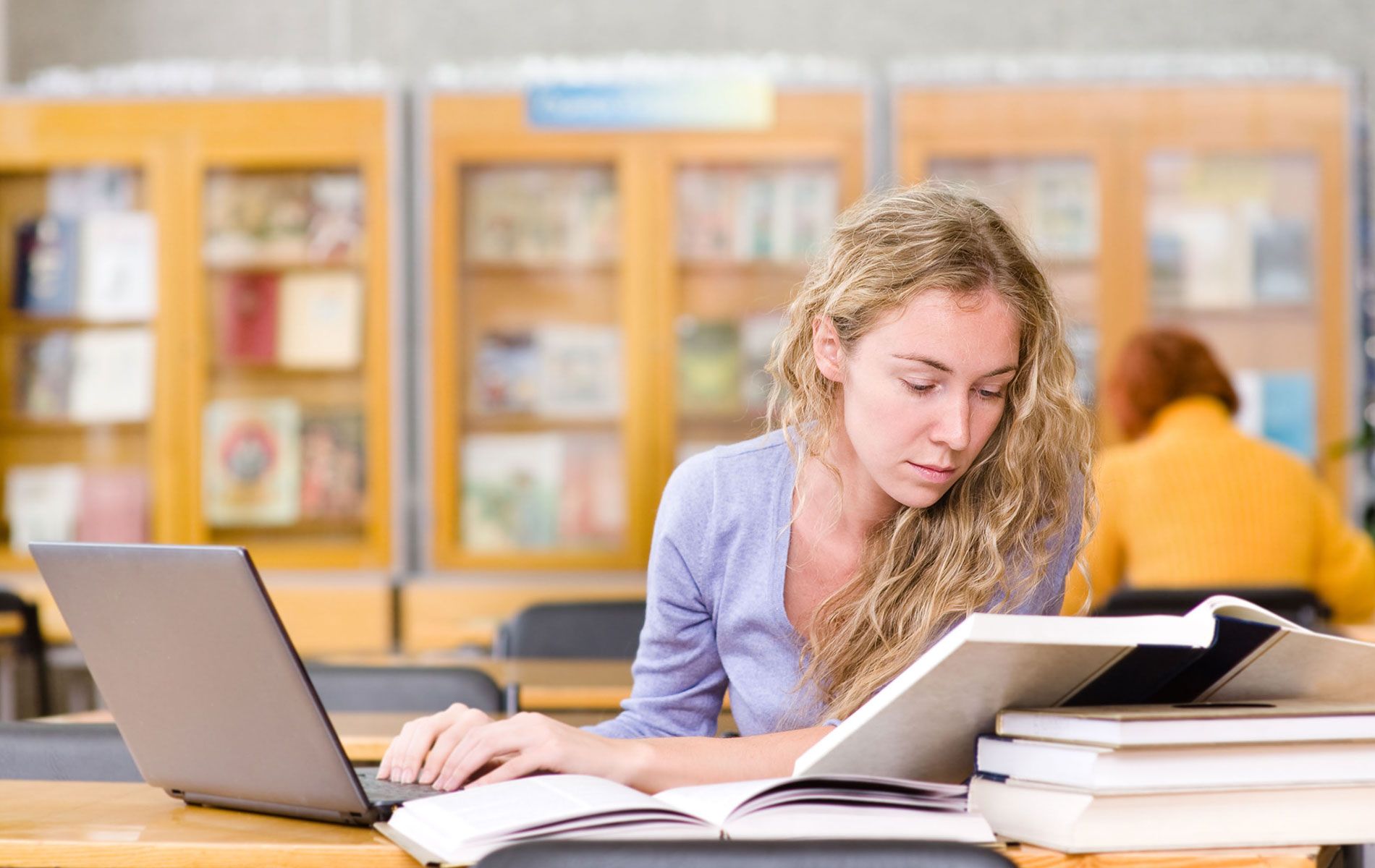 Blonde woman studies at desk in library with laptop, books. Shelves with books in background.
