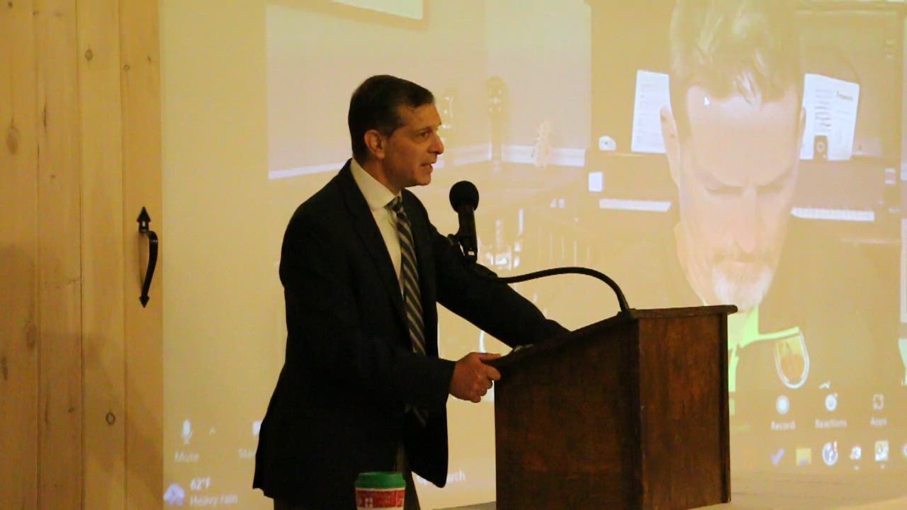 Man in suit speaks at a wooden podium with a screen behind him.
