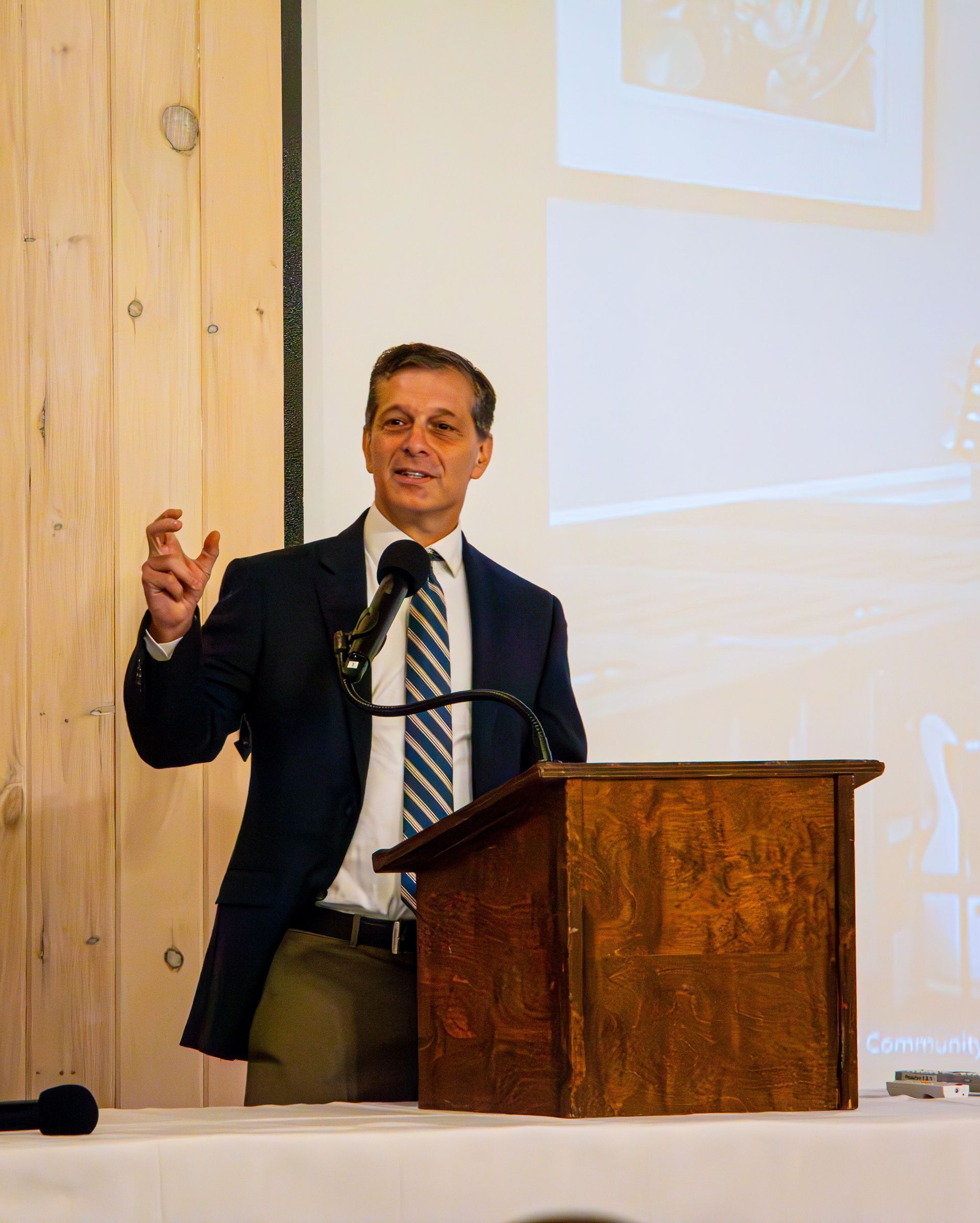 Man speaking at a podium, gesturing with his hand. He wears a suit, standing in front of a light-colored wooden backdrop.