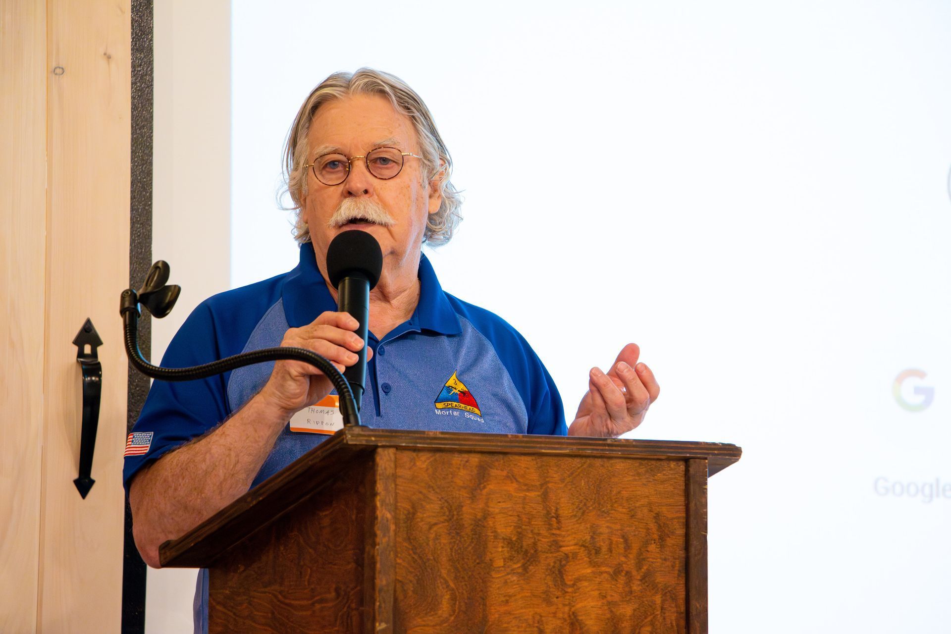 Man with gray hair and mustache speaks at a podium, holding a microphone. Blue shirt. Blank white screen behind him.