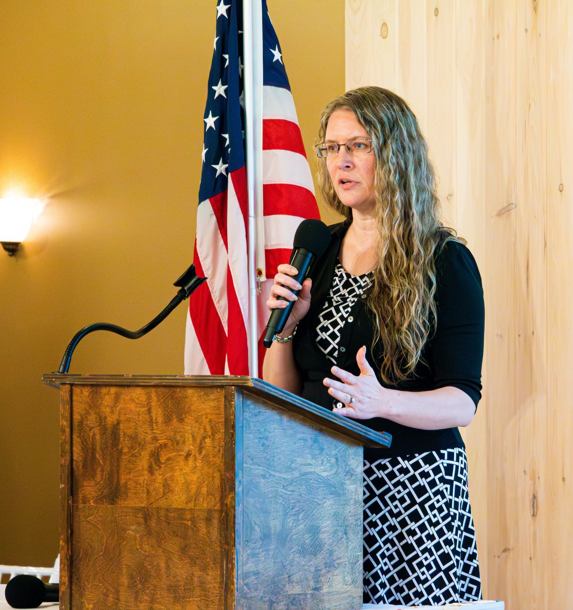 Woman speaking at a podium, holding a microphone. US flag in background.
