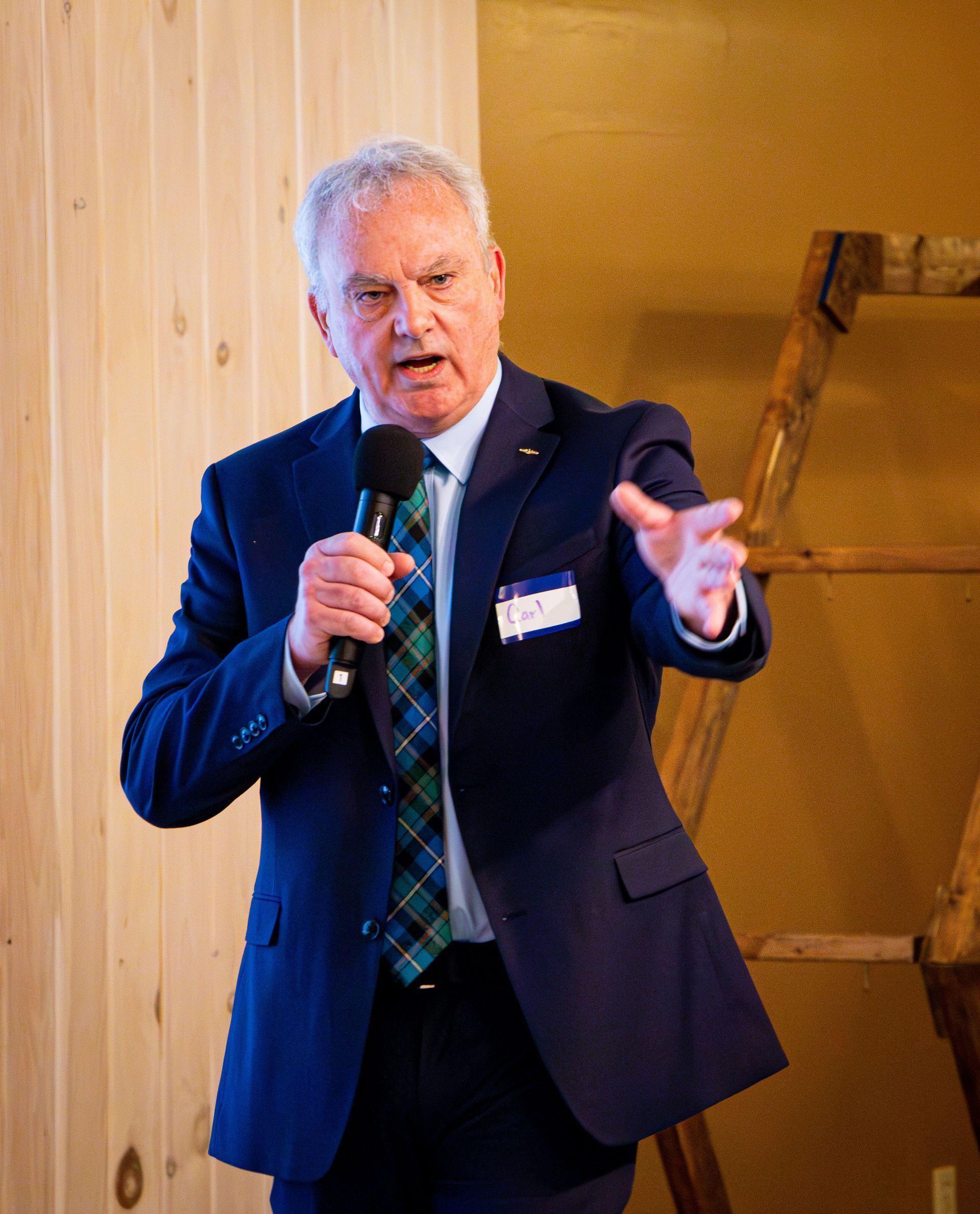 Man in a blue suit speaks into a microphone, gesturing with his right hand, standing indoors.