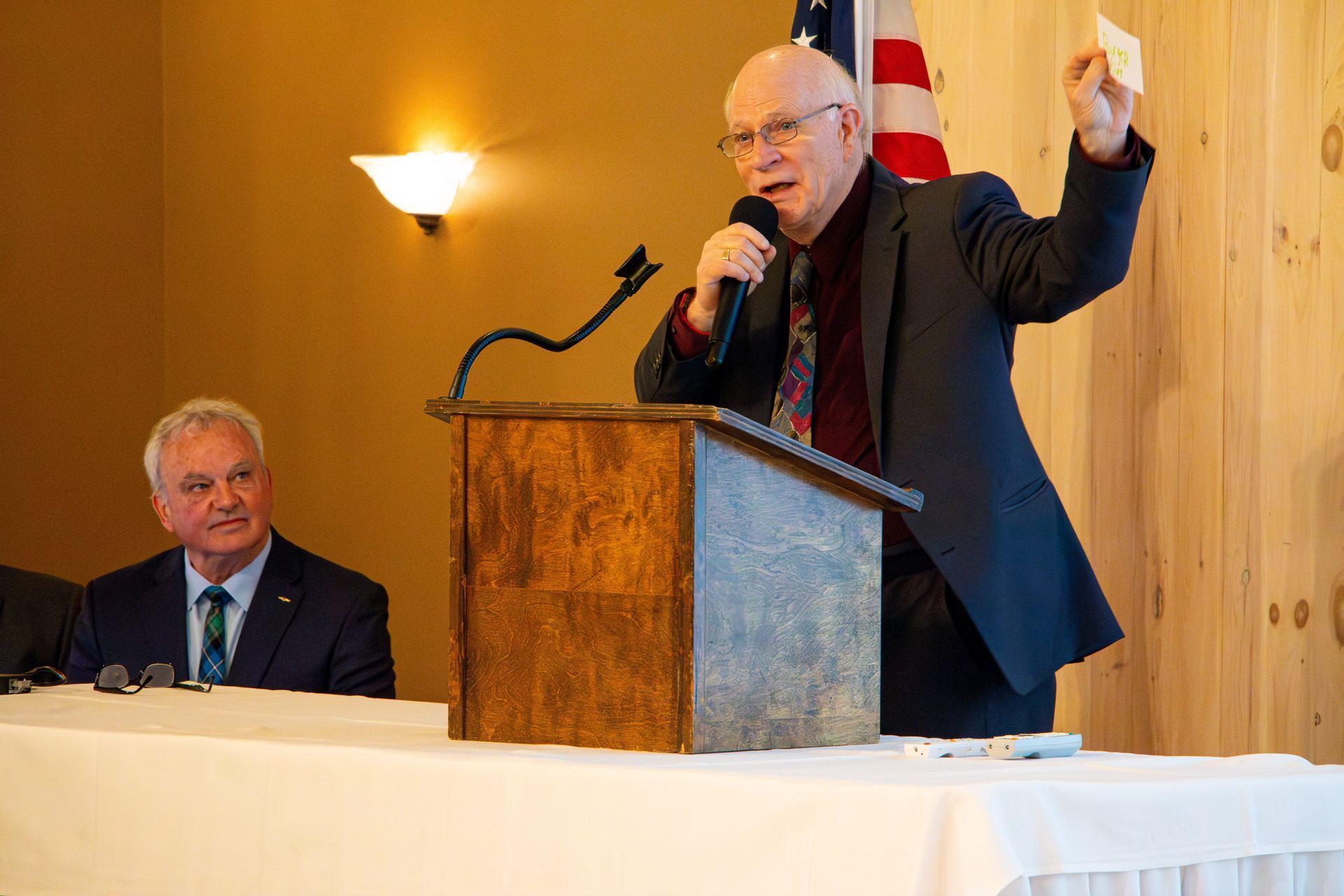 Man speaking at podium, holding card. Another man sits at a table. Room has American flag.