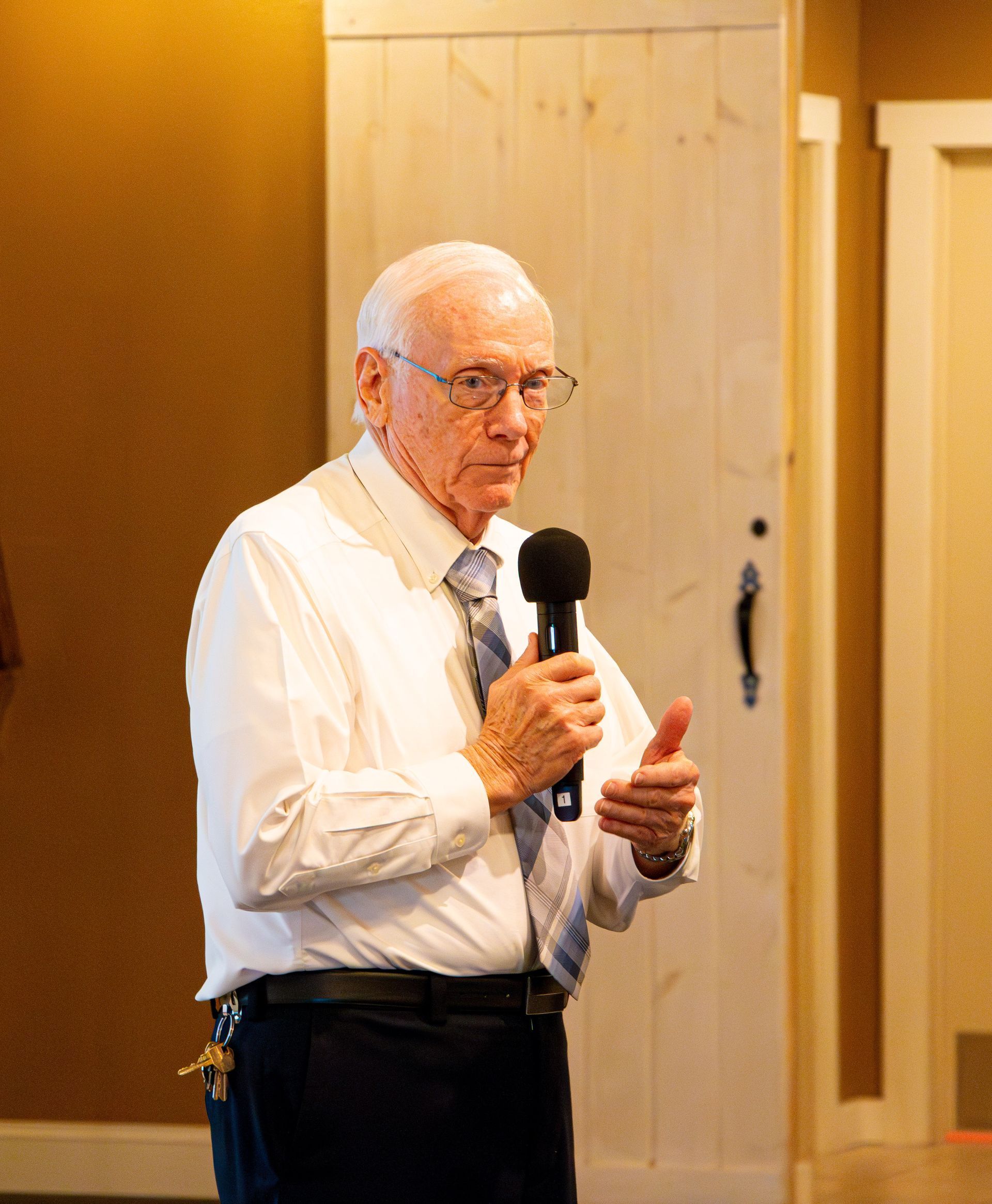 Older man speaking into a microphone, wearing a white shirt and tie. He stands in front of a light wooden door.