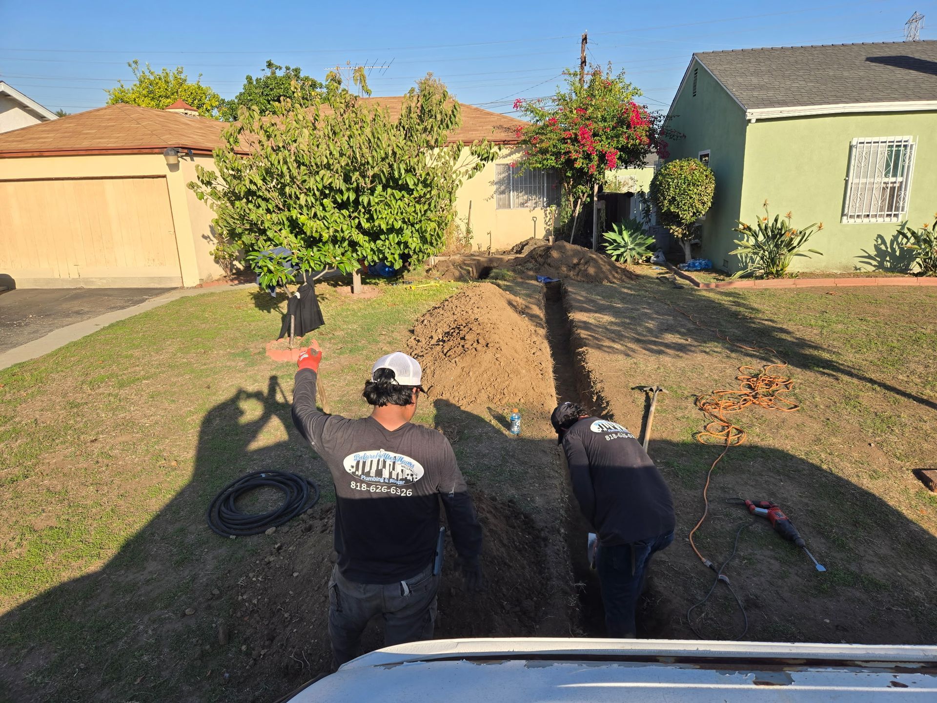 Two workers dig a long trench in a residential front yard on a sunny day.