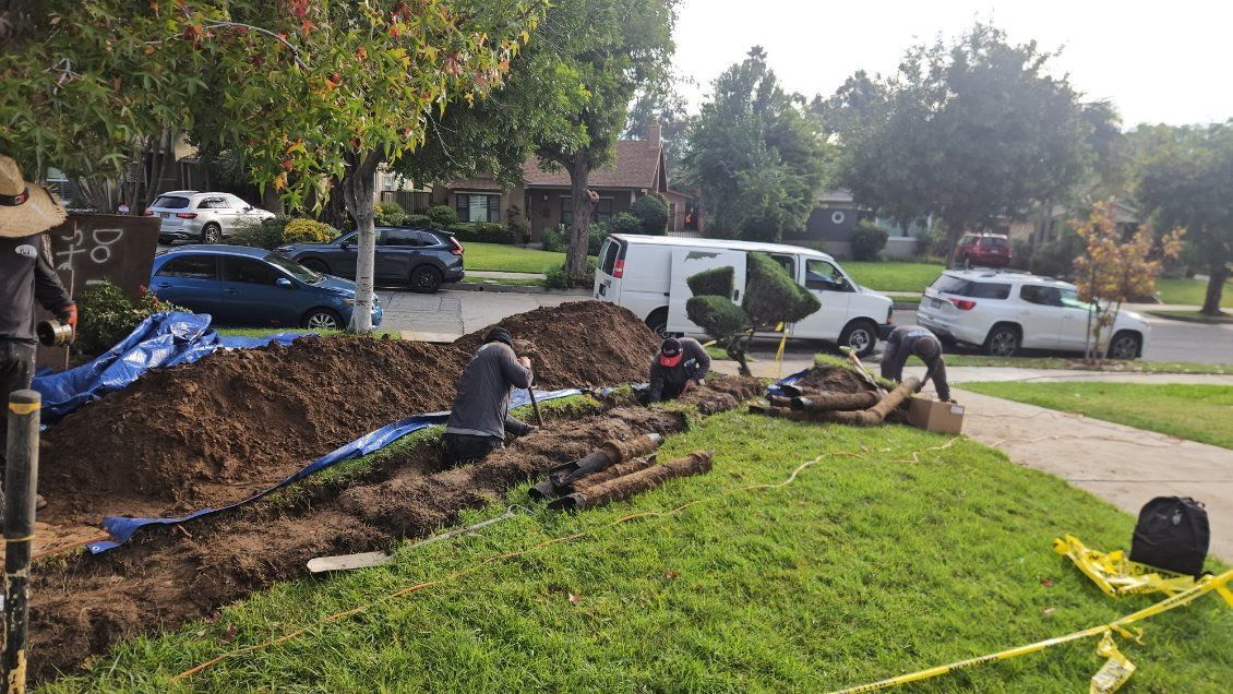 Workers dig a trench along a suburban street, with piles of dirt, equipment, and parked vehicles visible nearby.