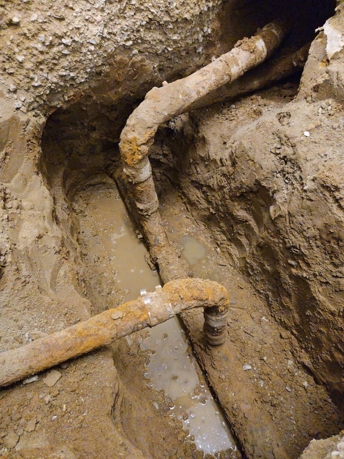 An overhead view of a muddy trench containing two old, rusted metal pipes with angled joints in the ground.