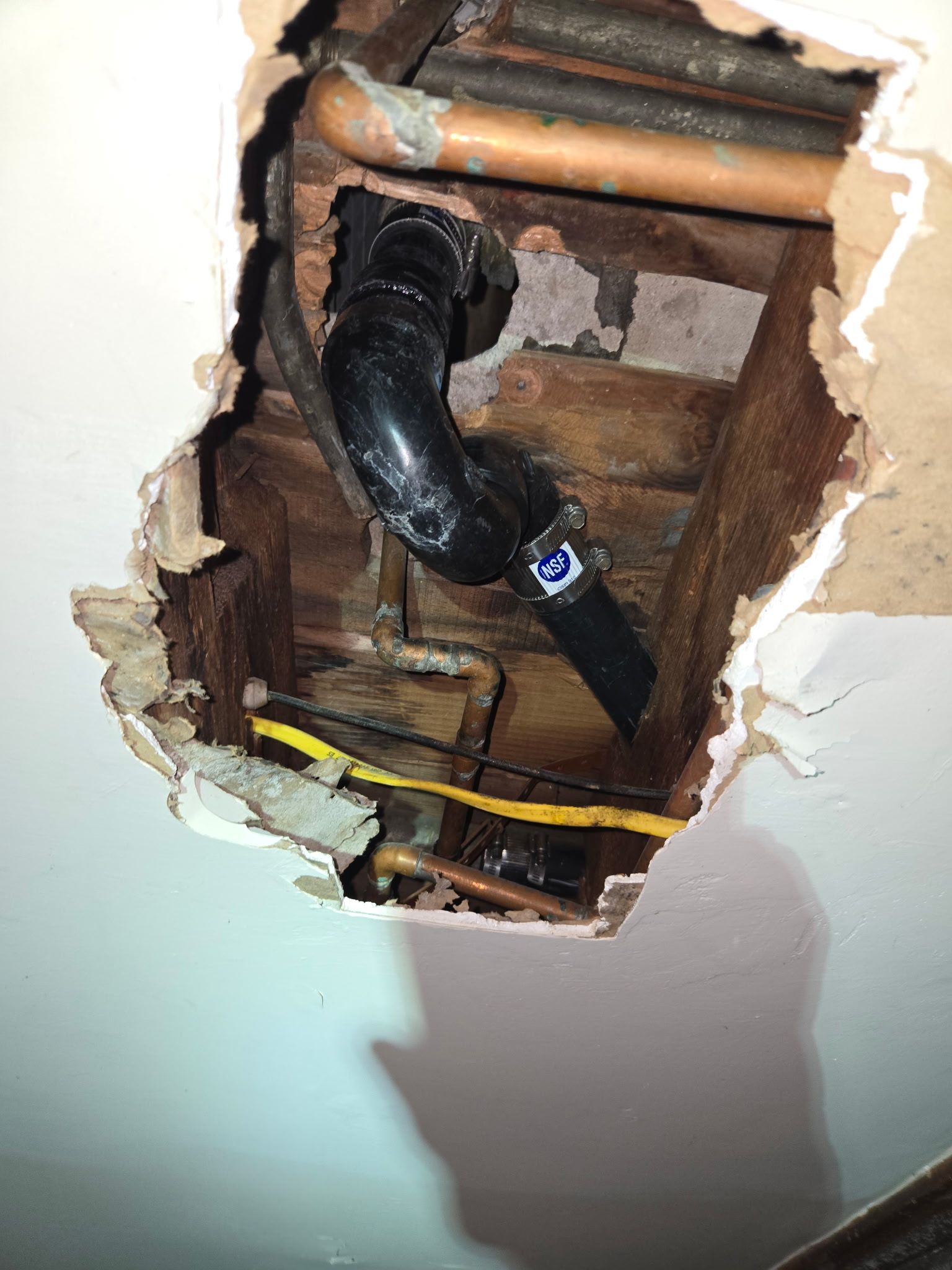 A view into an open wall hole showing black ABS plumbing pipes and copper water lines among wooden wall studs.