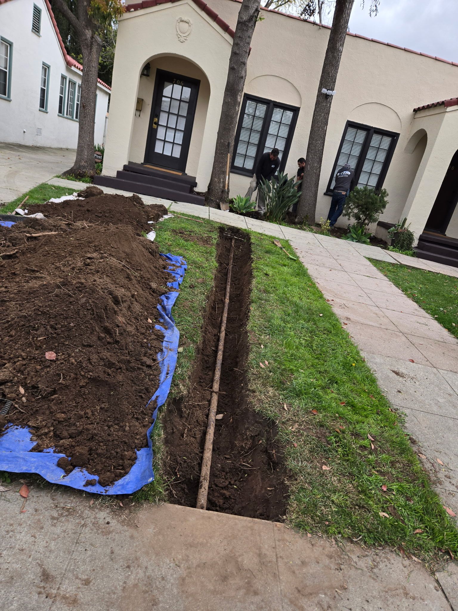 A trench is dug in a grassy front lawn next to a large pile of dirt on a blue tarp, with workers near a house entrance.