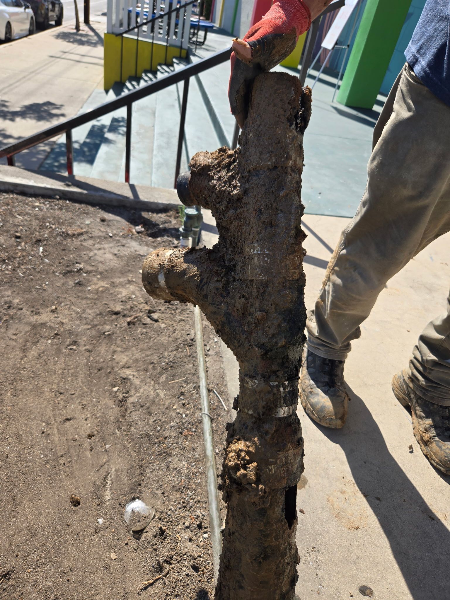 A person in work gloves holds a heavily corroded, mud-covered metal pipe section against an outdoor sidewalk background.