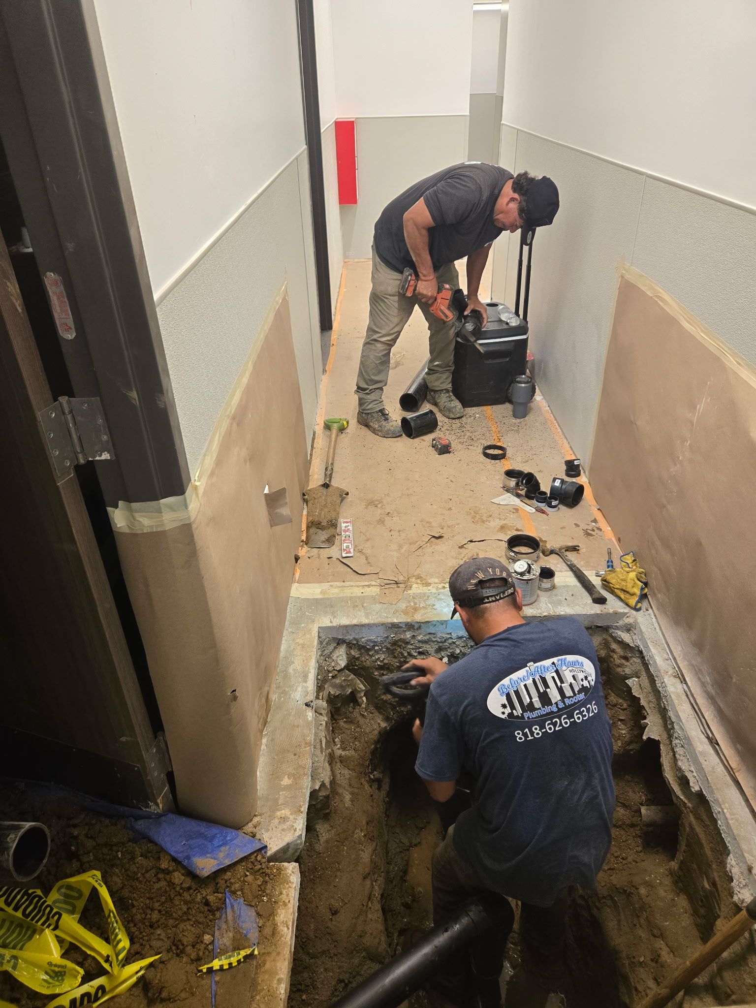 Two workers repair plumbing in a narrow hallway with a section of the concrete floor cut out to access pipes.