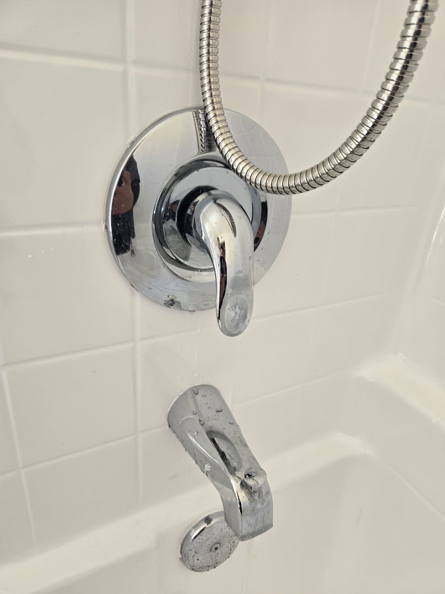 Chrome shower faucet and handle installed on white tiled wall above a bathtub.