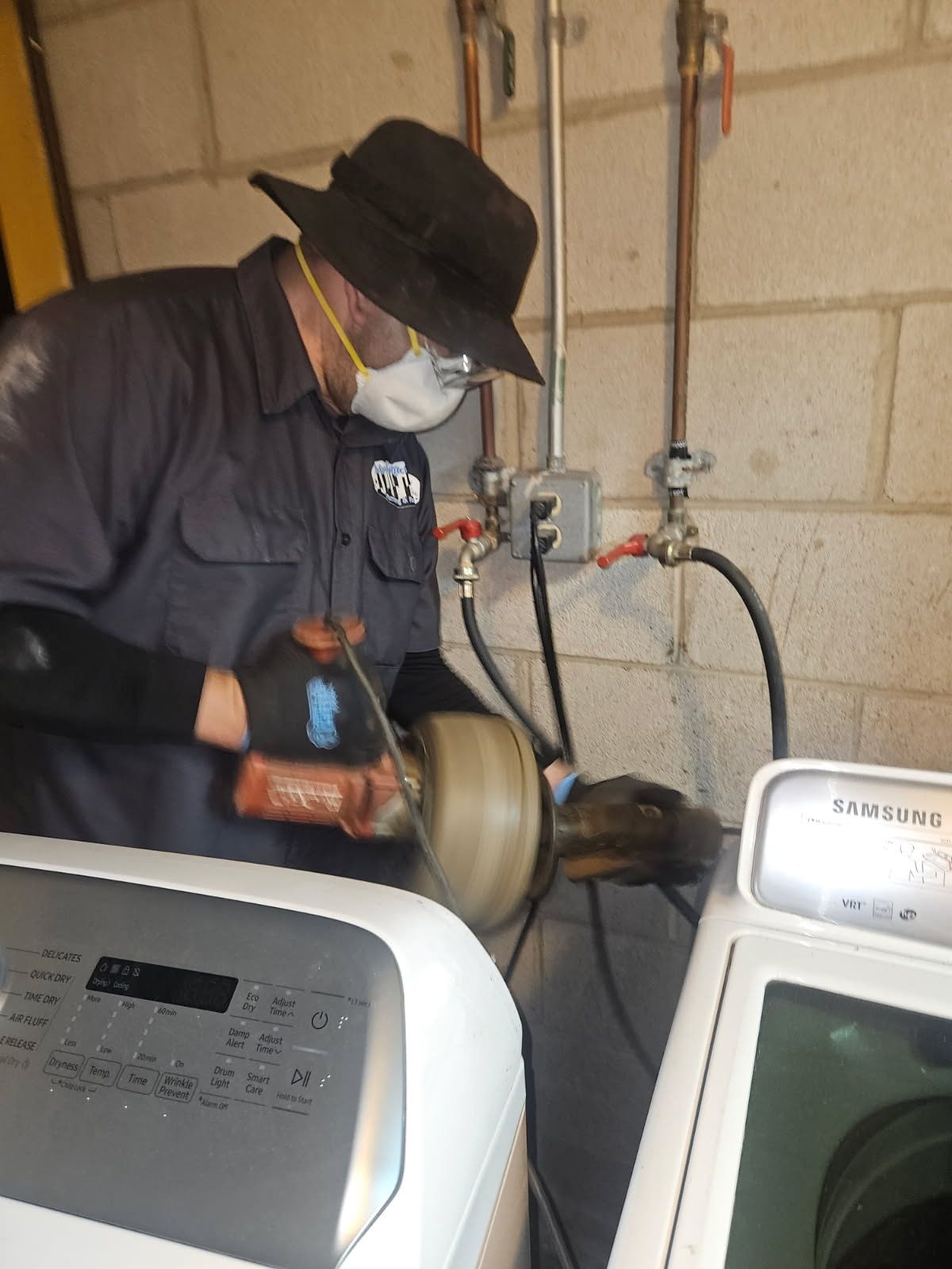 A person wearing a hat and respirator uses a power tool to cut a pipe behind two washing machines in a basement.