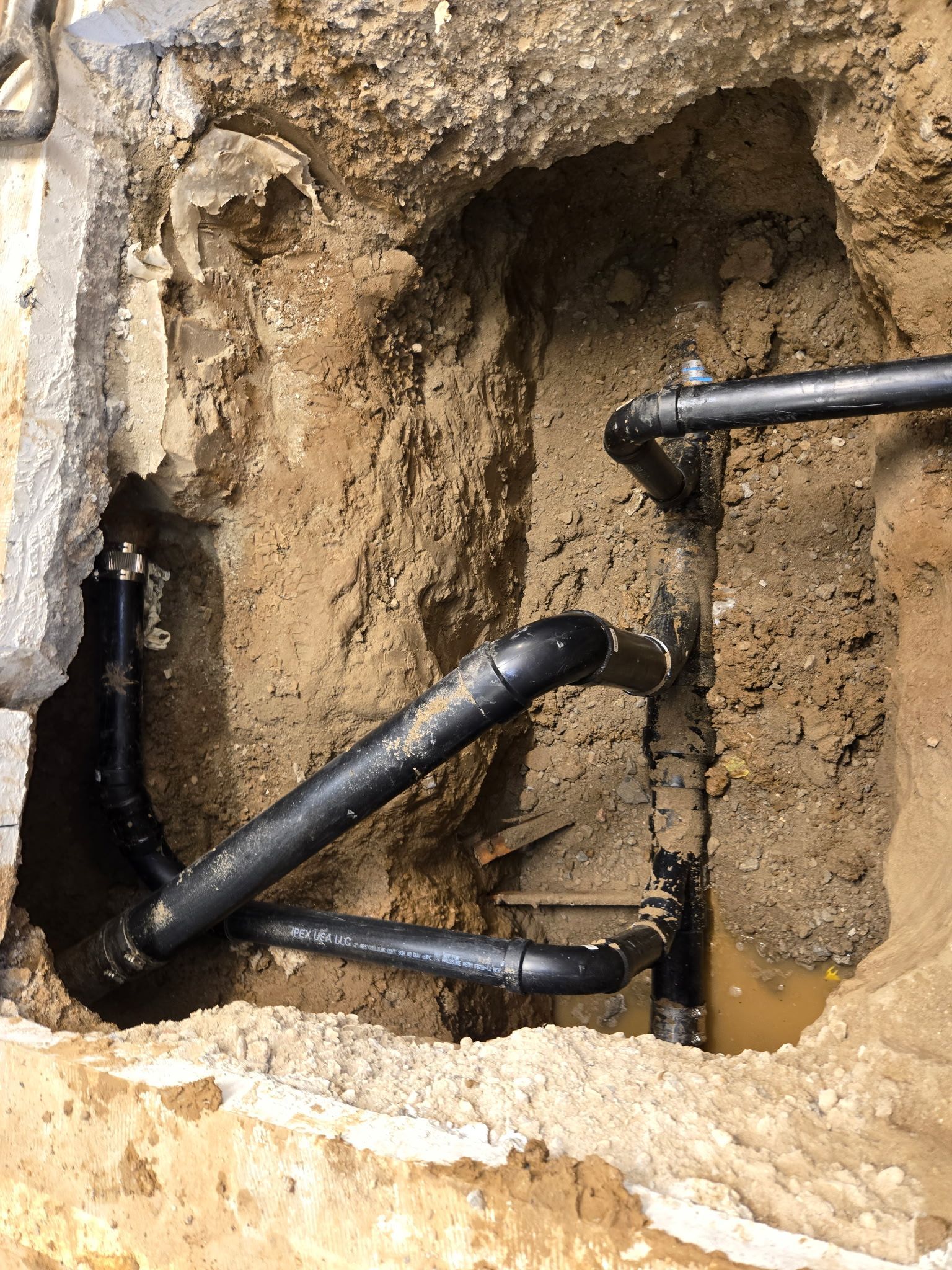 A square hole in the ground reveals a network of black PVC plumbing pipes connected with various fittings and joints.