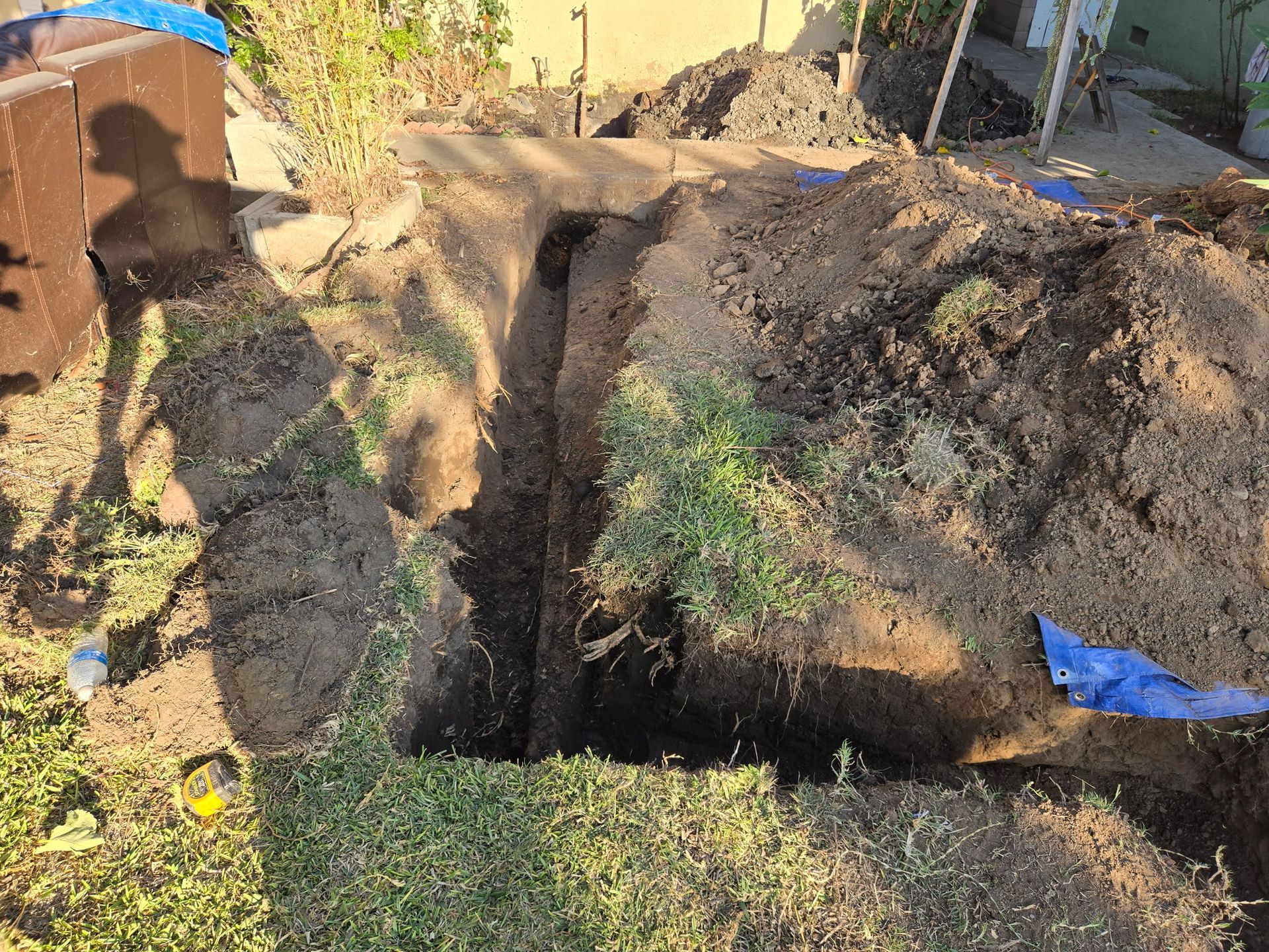 A deep, narrow trench dug into a backyard lawn, surrounded by piles of excavated dirt, with a blue tarp visible.