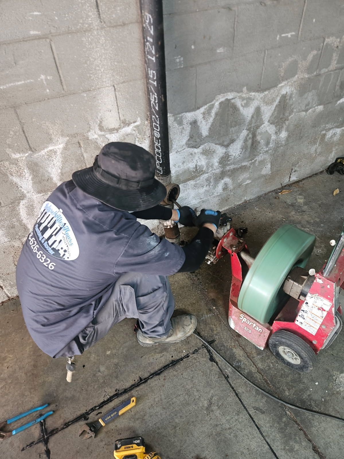 A technician in a uniform and hat kneels to operate a pipe cleaning machine against a concrete wall.