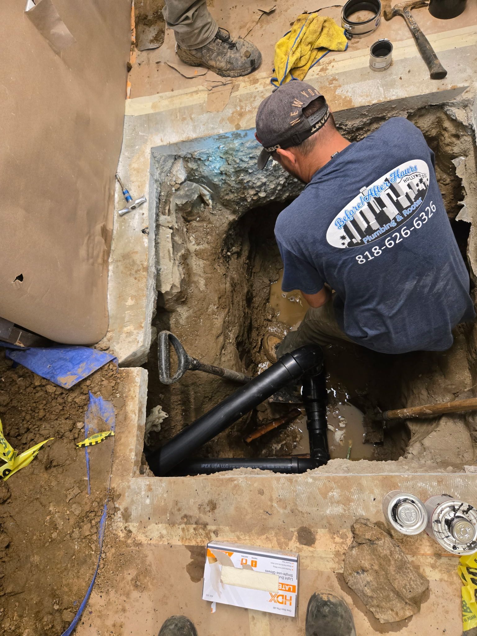 A worker in a blue shirt repairs underground pipes within a concrete-bordered excavation pit.