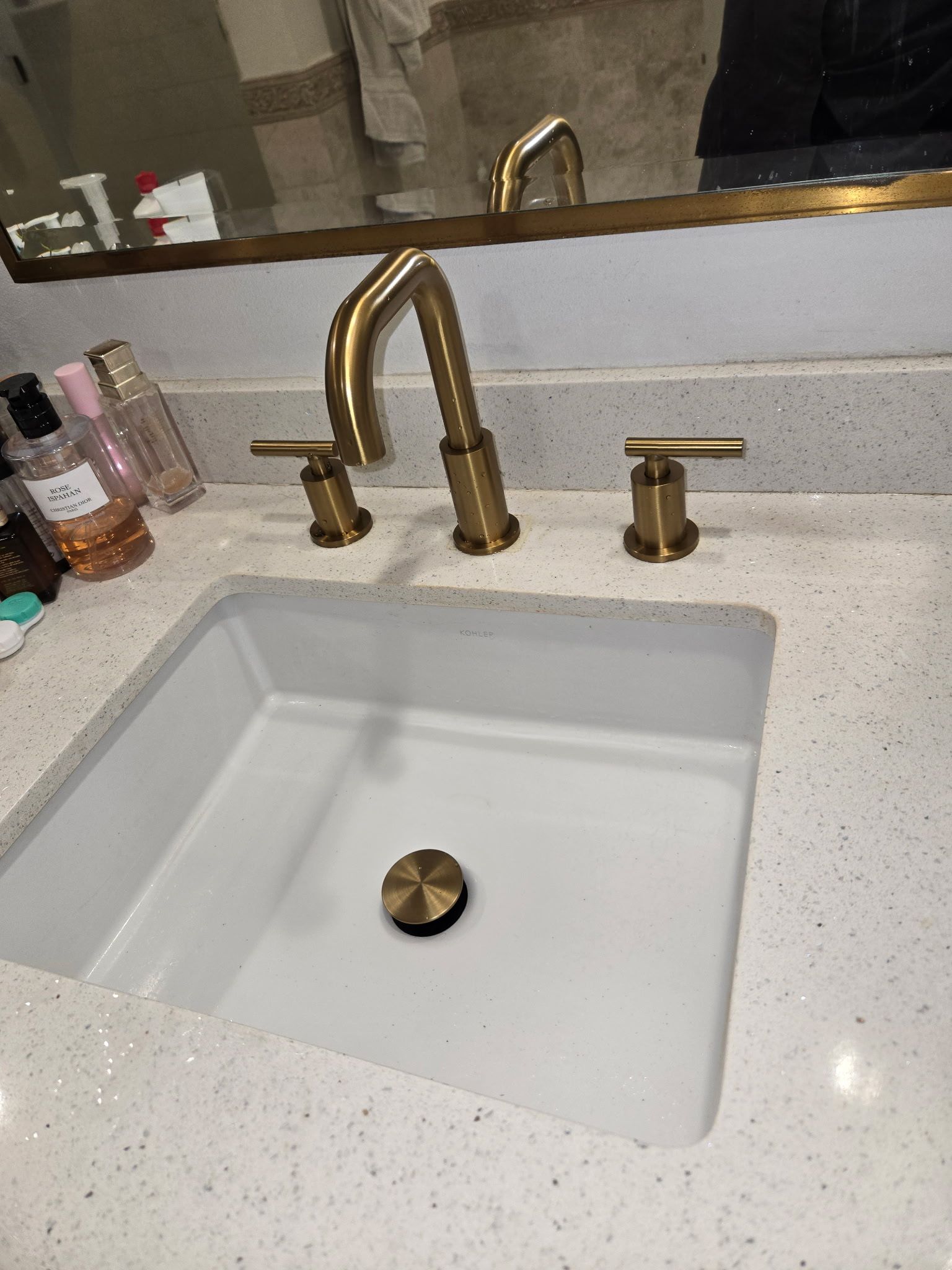 A square white bathroom sink with a brushed gold faucet and handles set into a white speckled quartz countertop.