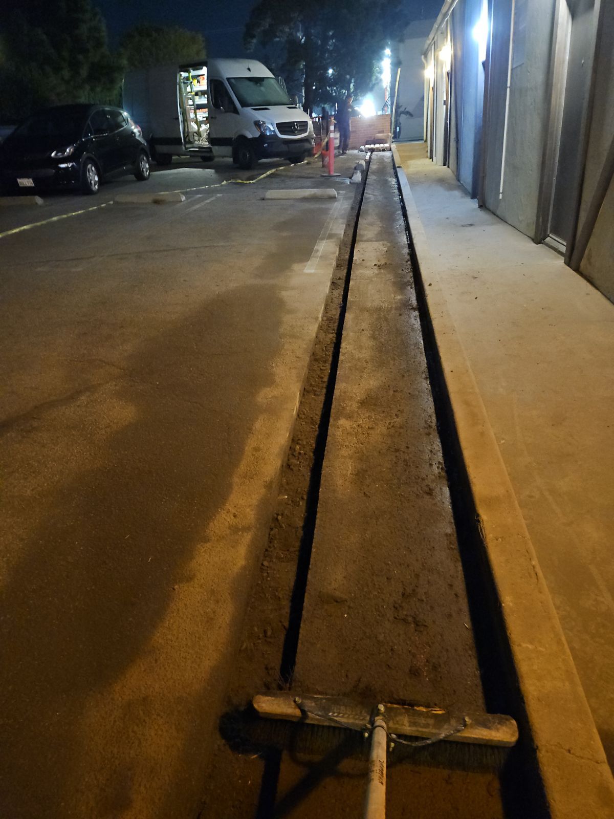 At night, a person uses a long-handled tool to smooth a freshly poured, narrow strip of concrete in a parking lot.