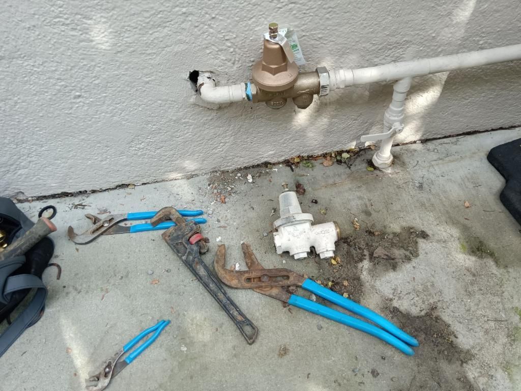 Plumbing tools and a disconnected valve assembly on concrete below a wall-mounted water pressure regulator.