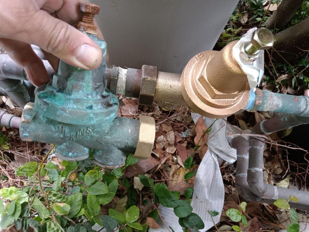 A hand holding an old, corroded brass water pressure regulator next to a newly installed one among outdoor foliage.