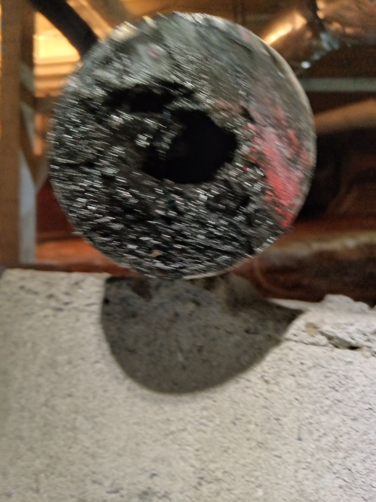 A close-up of a damaged, dark gray circular pipe showing heavy corrosion and a hole, set against a blurry background.