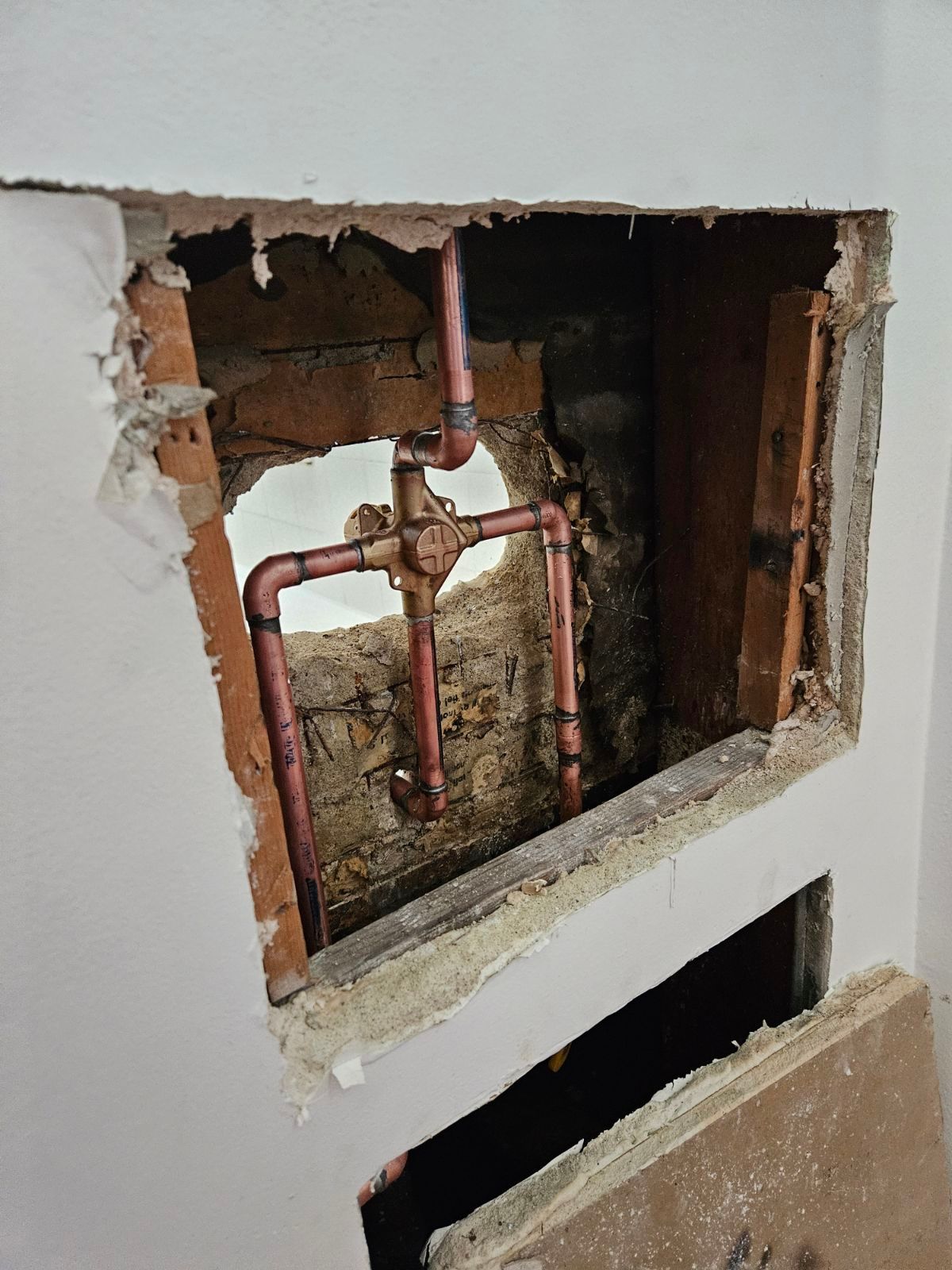 A rectangular hole in a white wall reveals copper plumbing pipes connected to a shower valve inside a wooden wall frame.